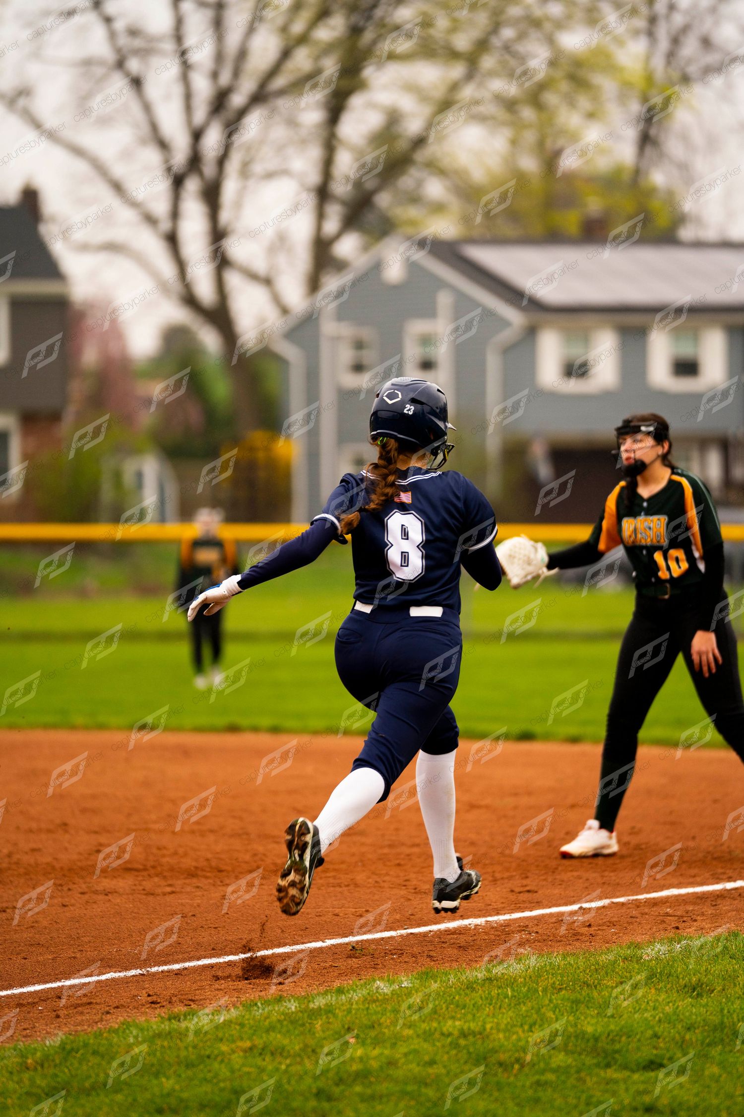 Softball player number 8 in navy uniform rounds third base as opposing fielder watches during a game.
