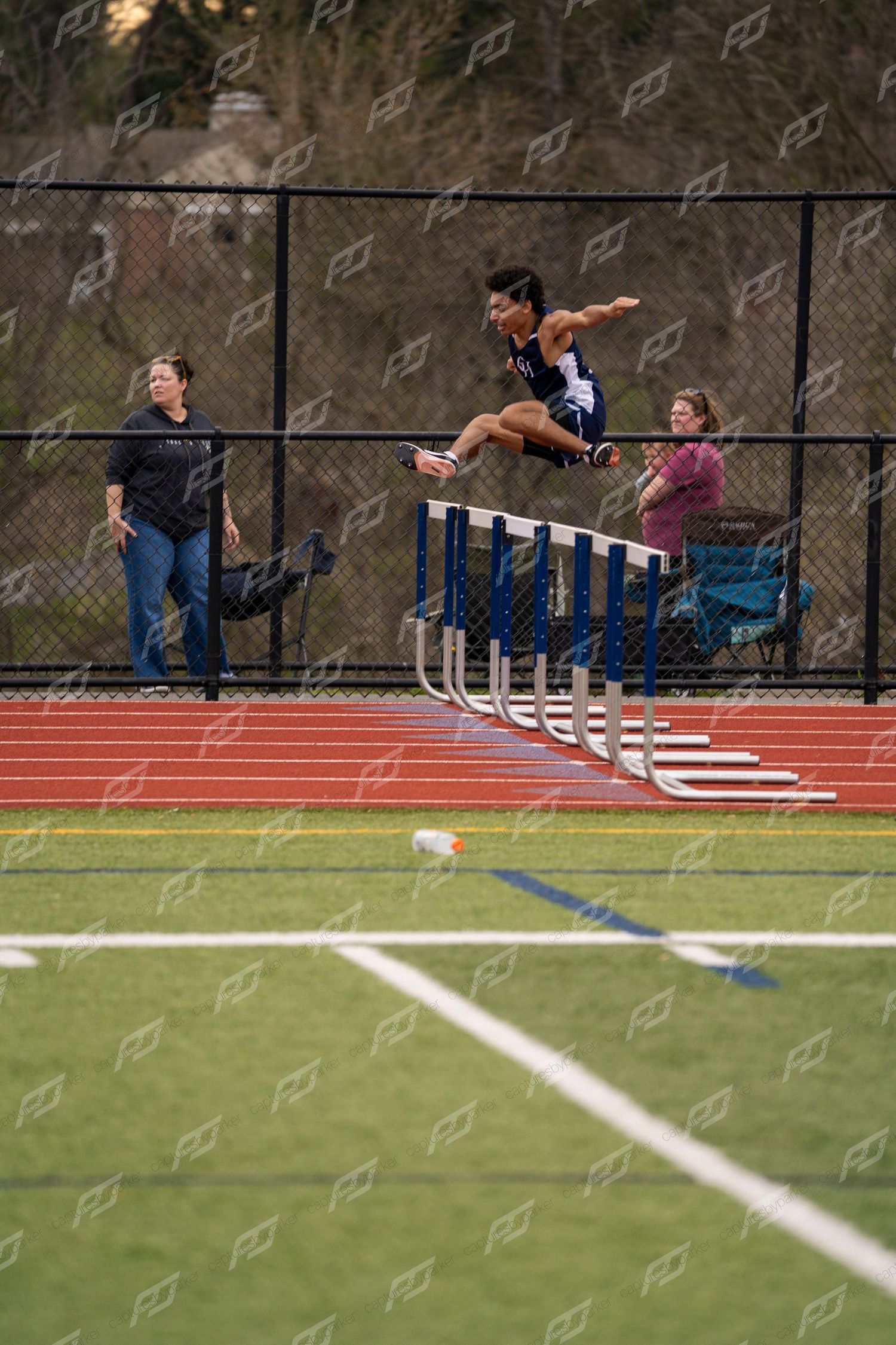 A track athlete leaps over hurdles during a high school track and field meet on a red track.