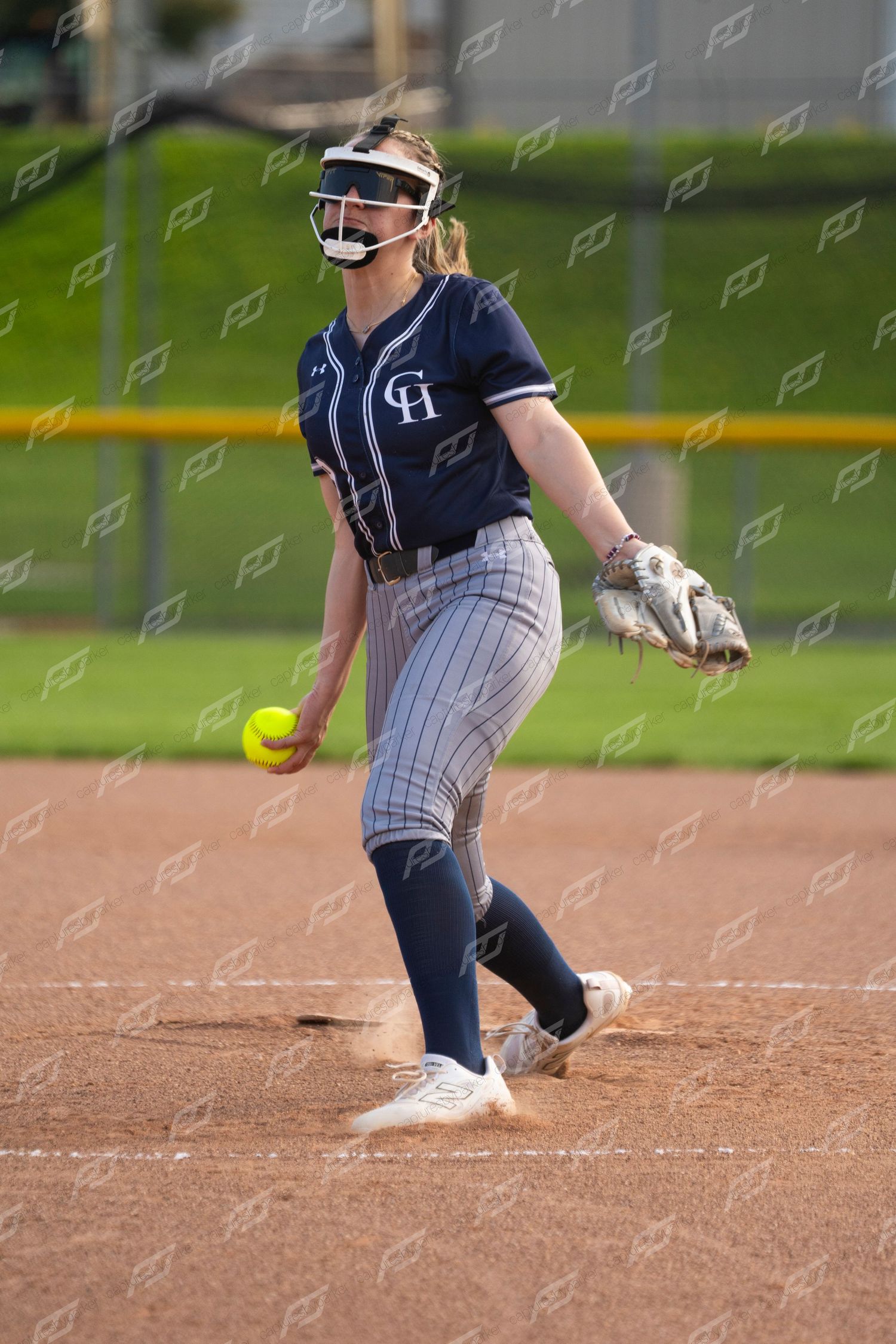 Female softball pitcher in navy CH uniform winds up to throw a yellow ball during a game.