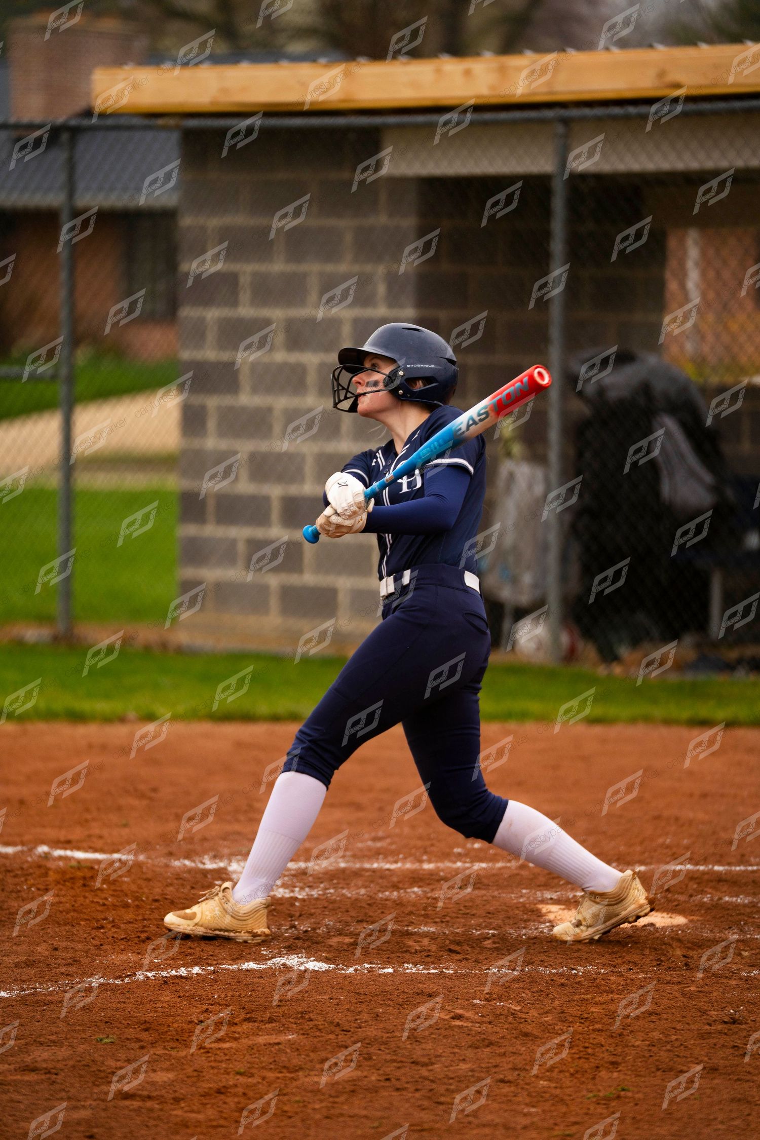 Female softball player in navy uniform follows through on a powerful swing during a game on a dirt field.