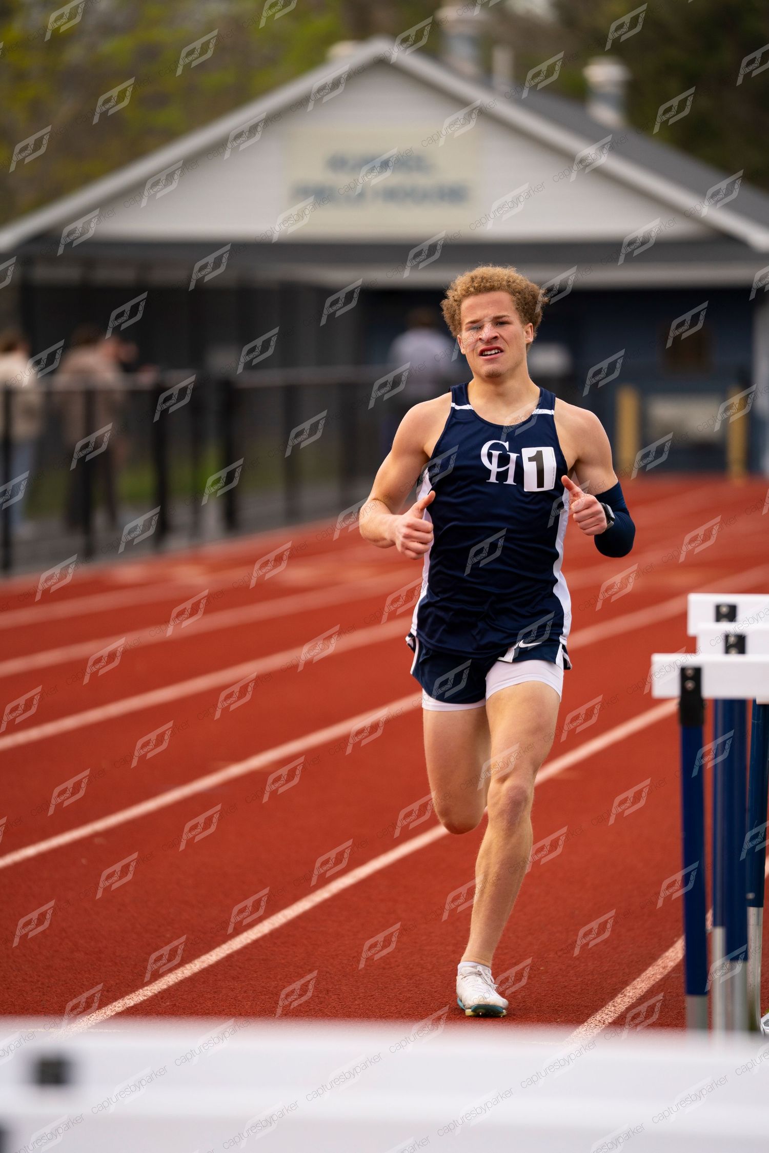 Male track athlete in navy CH uniform running past hurdles on red track.