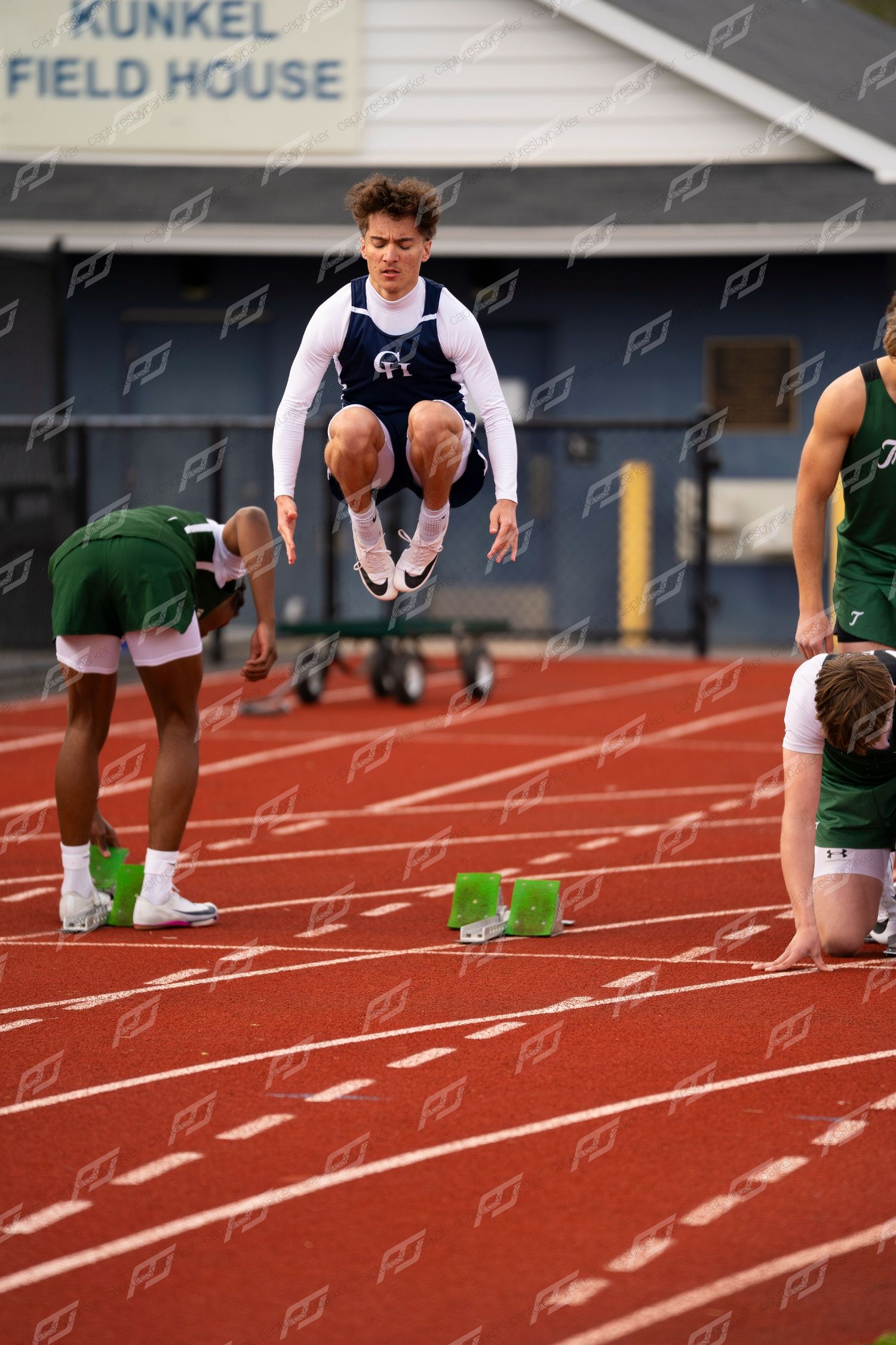 Male track athlete in navy uniform leaps at starting blocks during outdoor meet at Kunkel Field House.