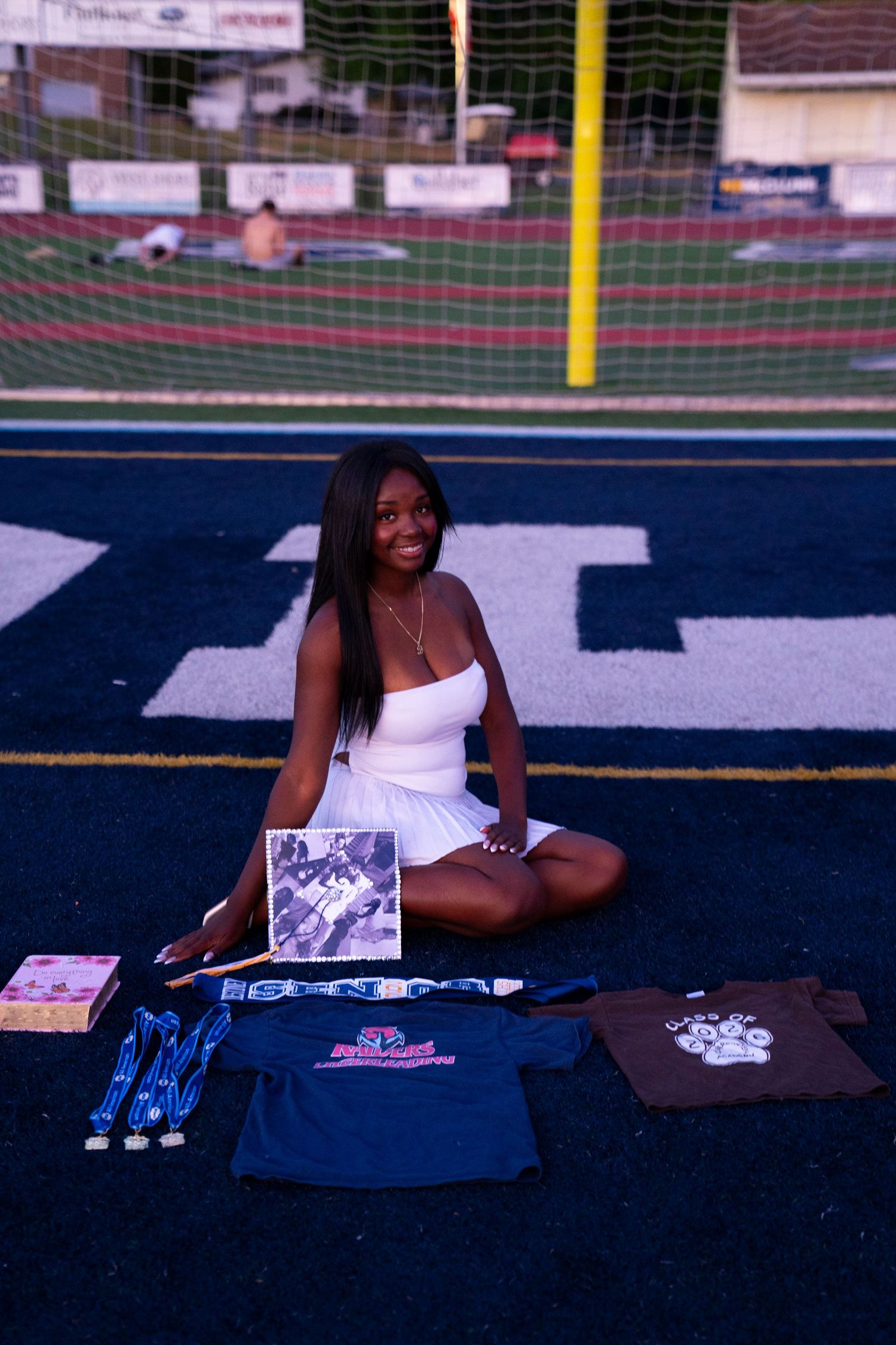 Young woman in white dress sits on track field surrounded by athletic awards and memorabilia.