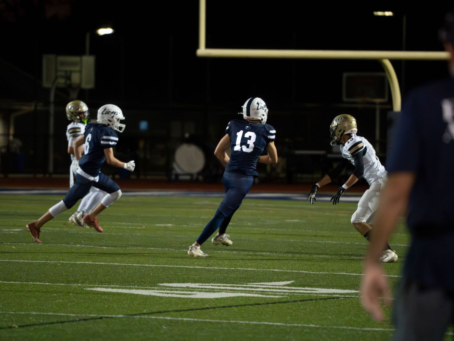 High school football player number 13 runs a route during a night game with defenders in pursuit on a lit field.