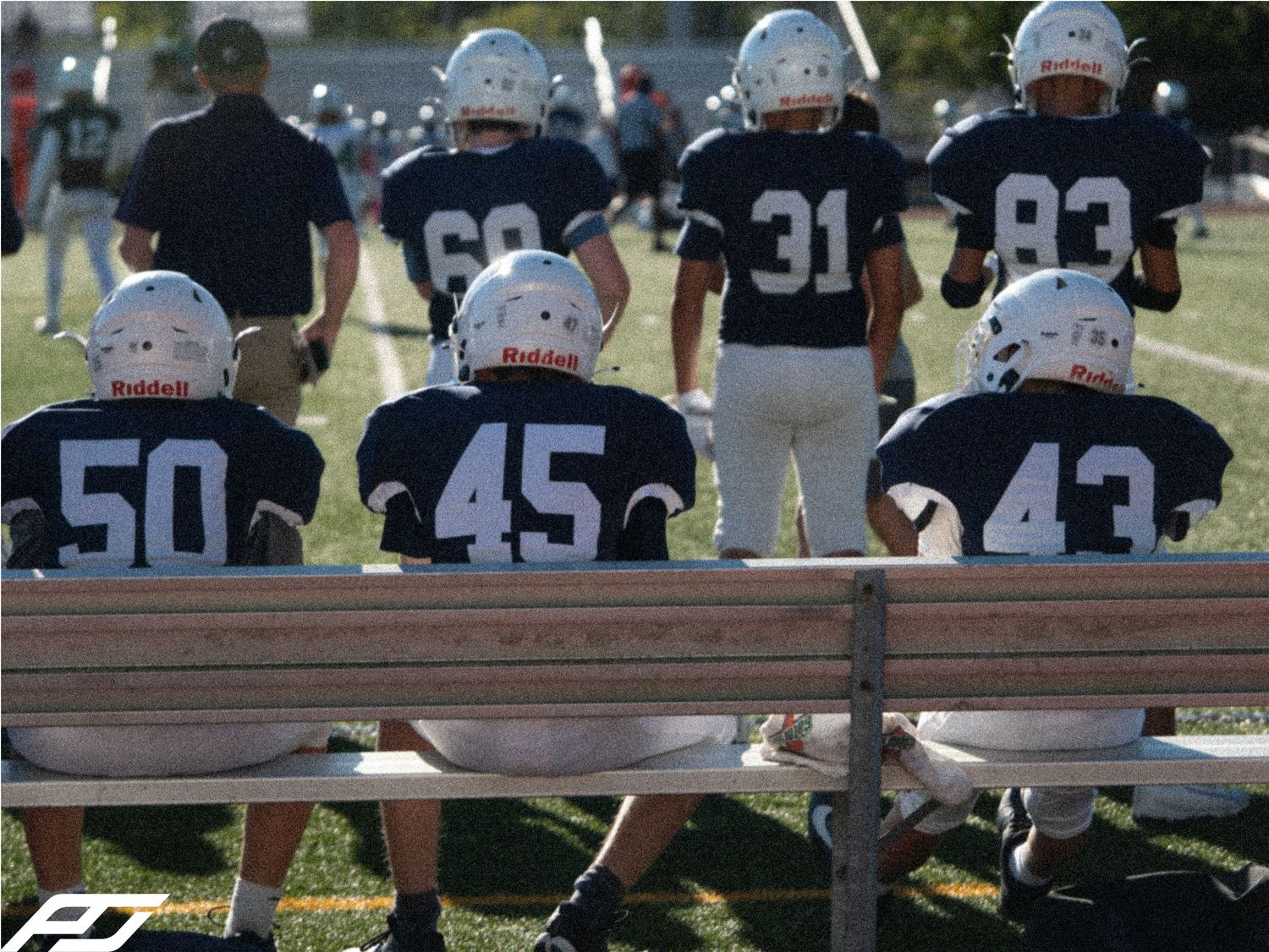 Youth football players in navy uniforms sit on a bench watching a game.