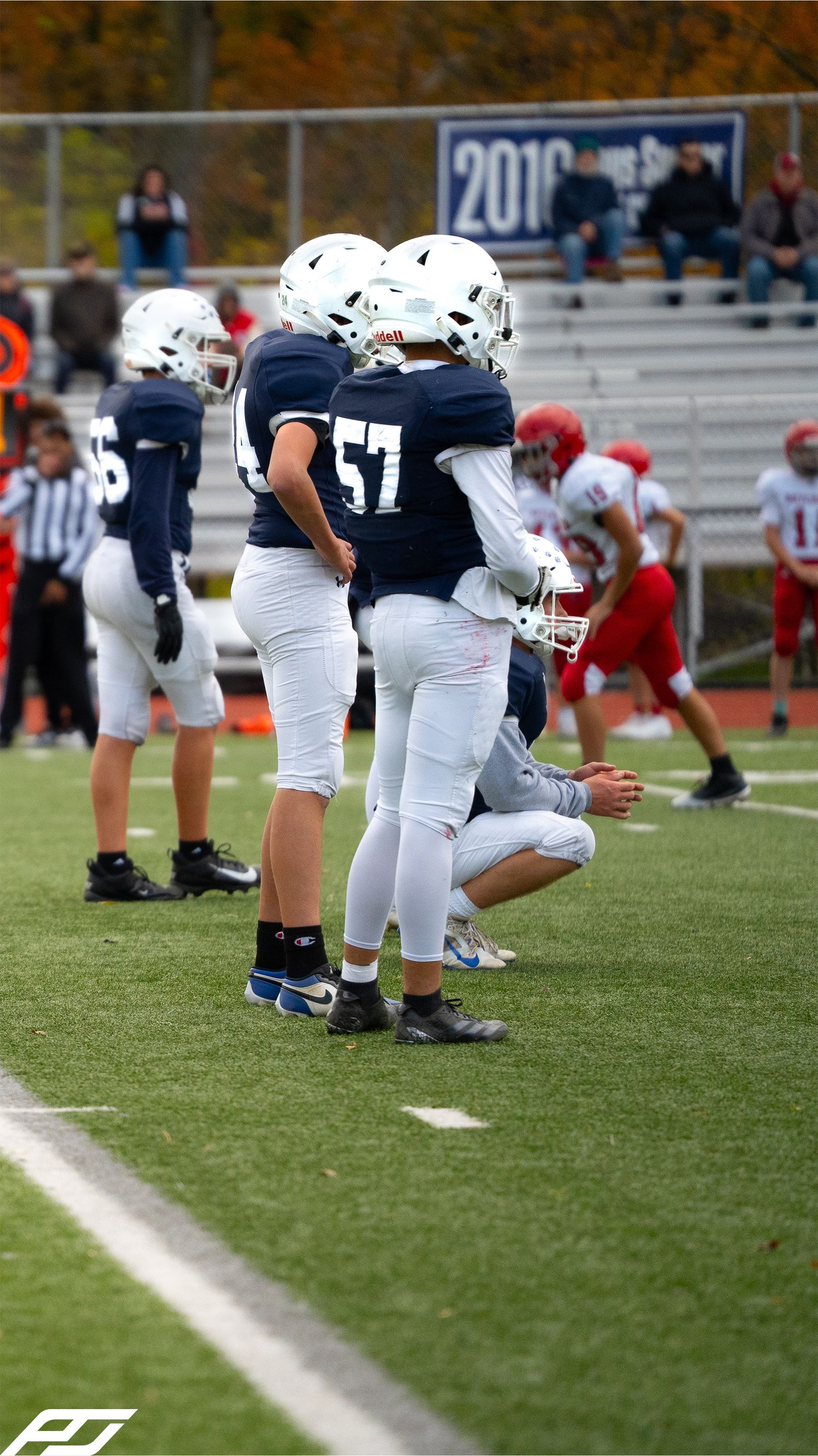 Youth football players in navy blue uniforms including number 57 huddle on the sideline during a fall game.