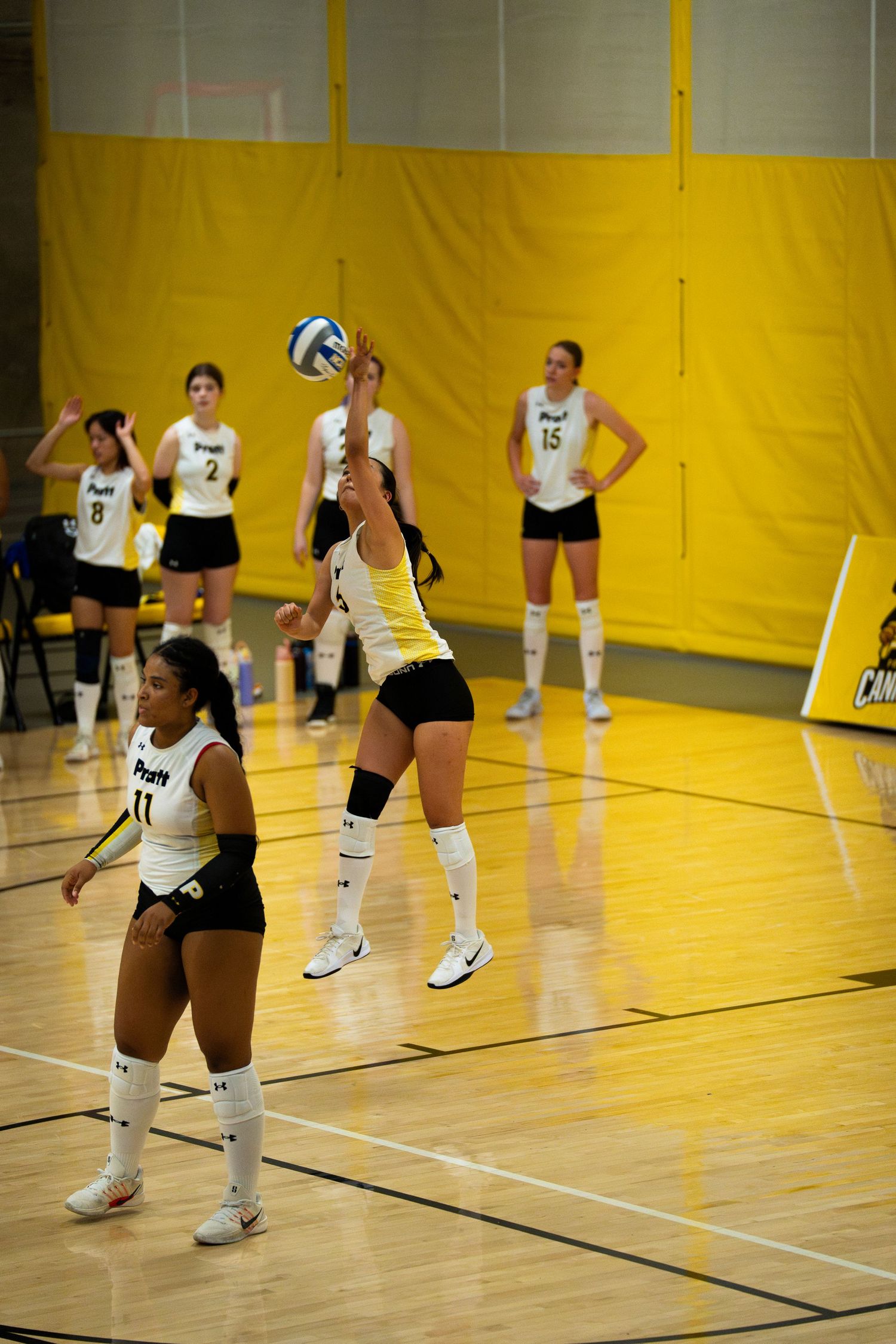 Female volleyball players in white and black uniforms compete indoors on a hardwood court with yellow walls.