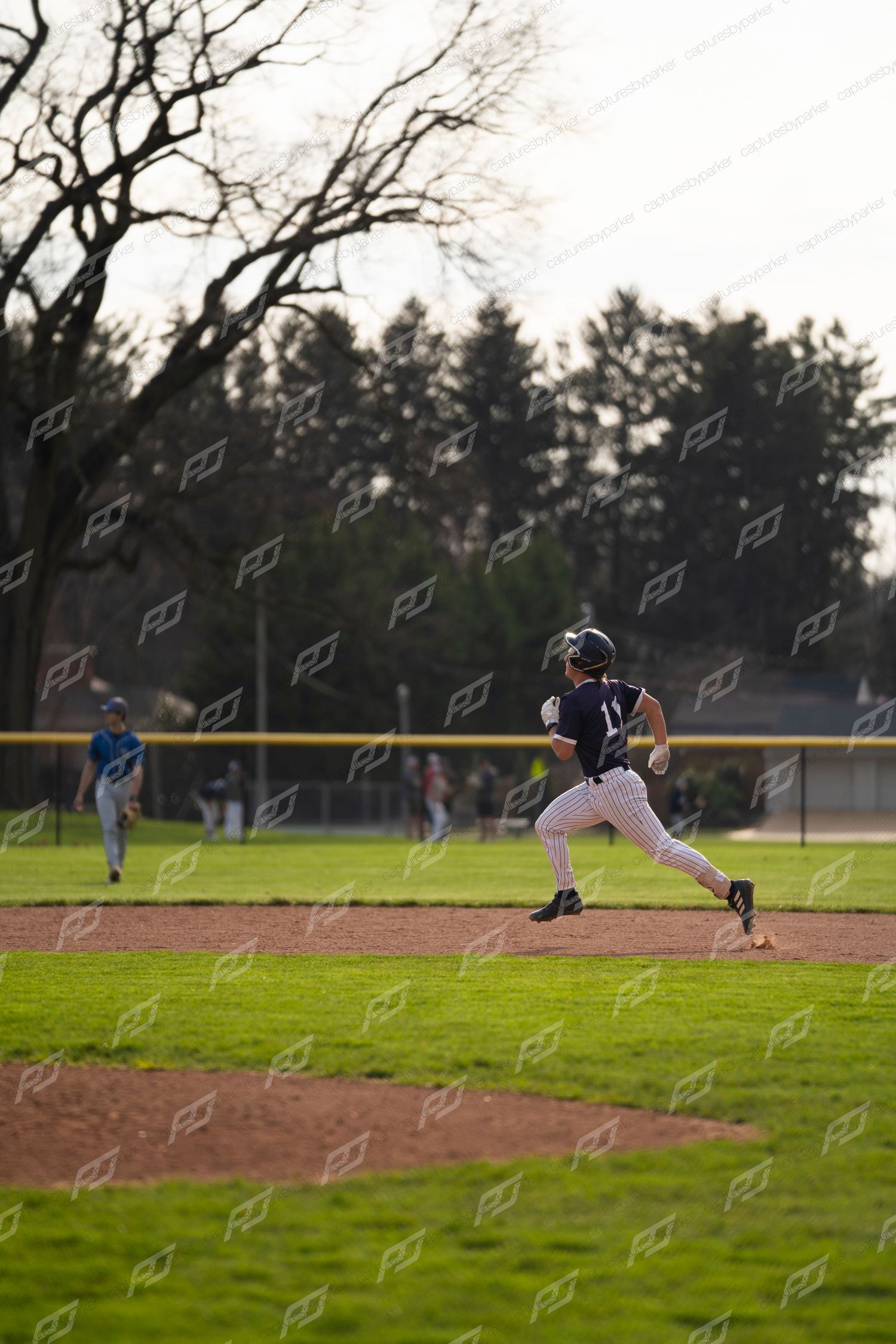 Baseball player rounds the bases during a game on a green field with trees in the background.
