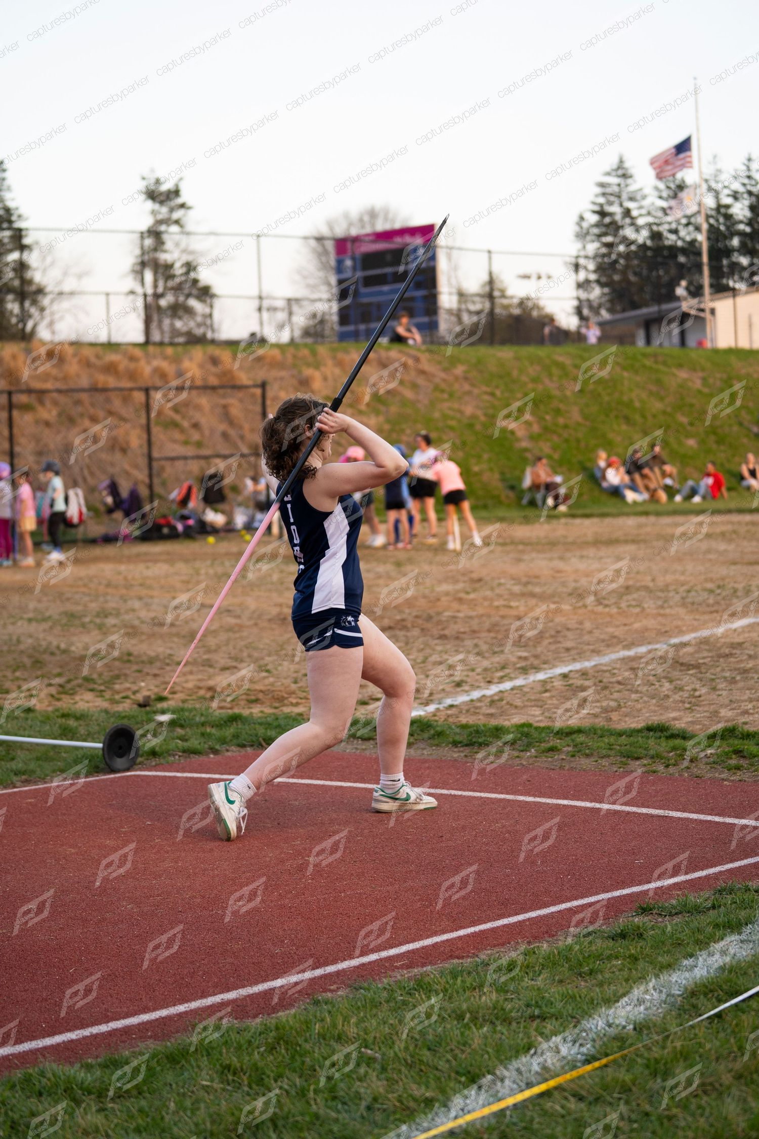 Female athlete releases javelin during outdoor high school track and field competition at dusk.