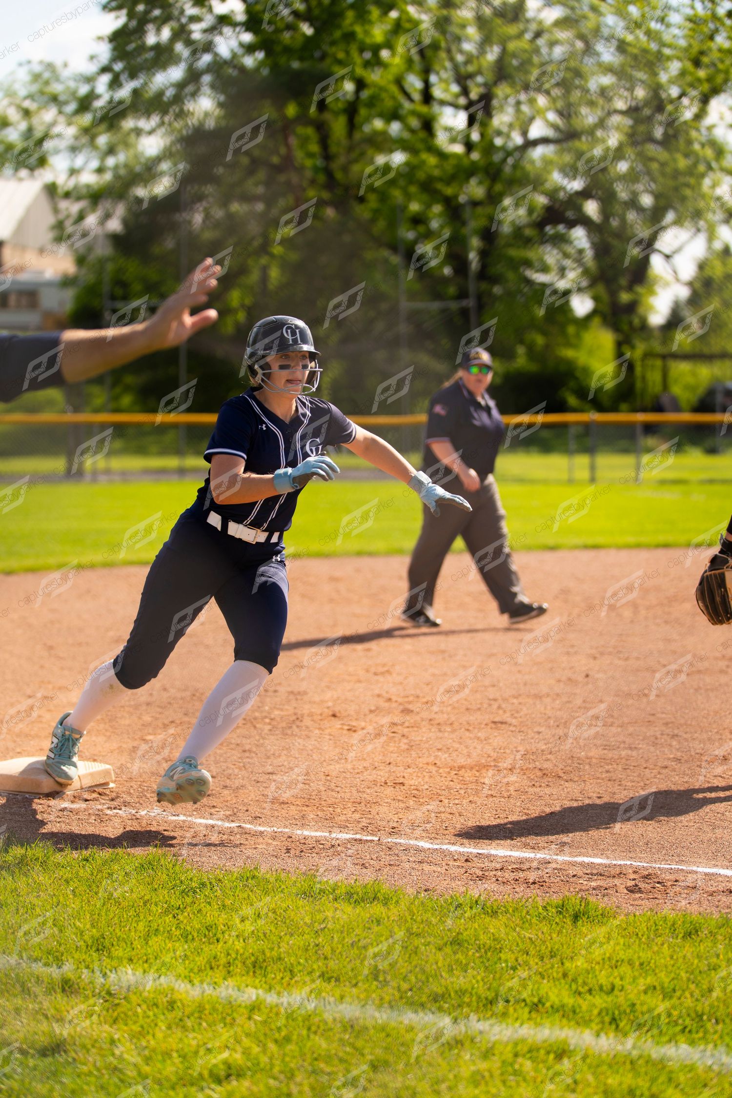 Softball player in navy uniform rounds third base while a coach signals during a game.