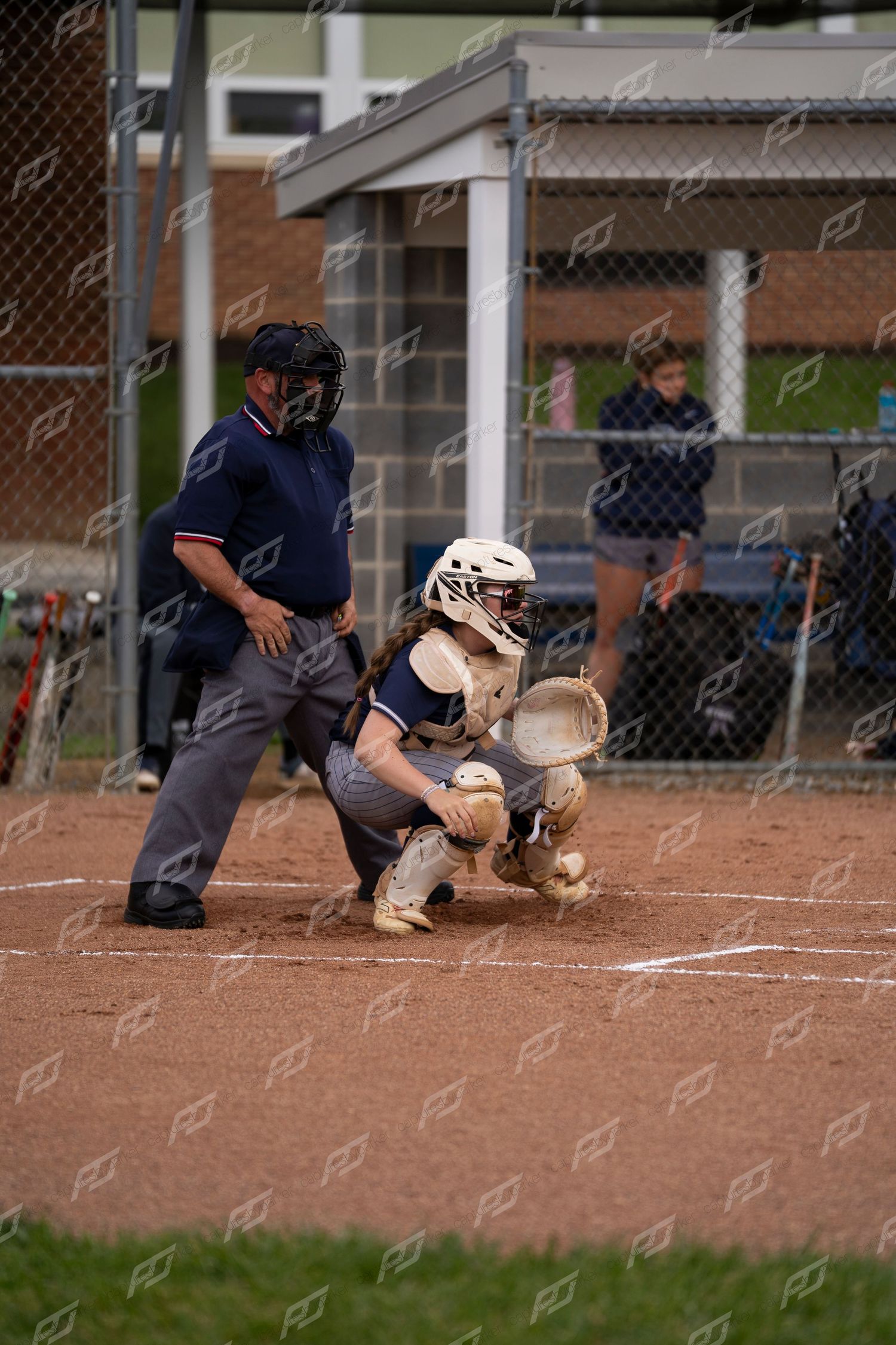 Softball catcher in full gear crouching at home plate with umpire positioned behind her.
