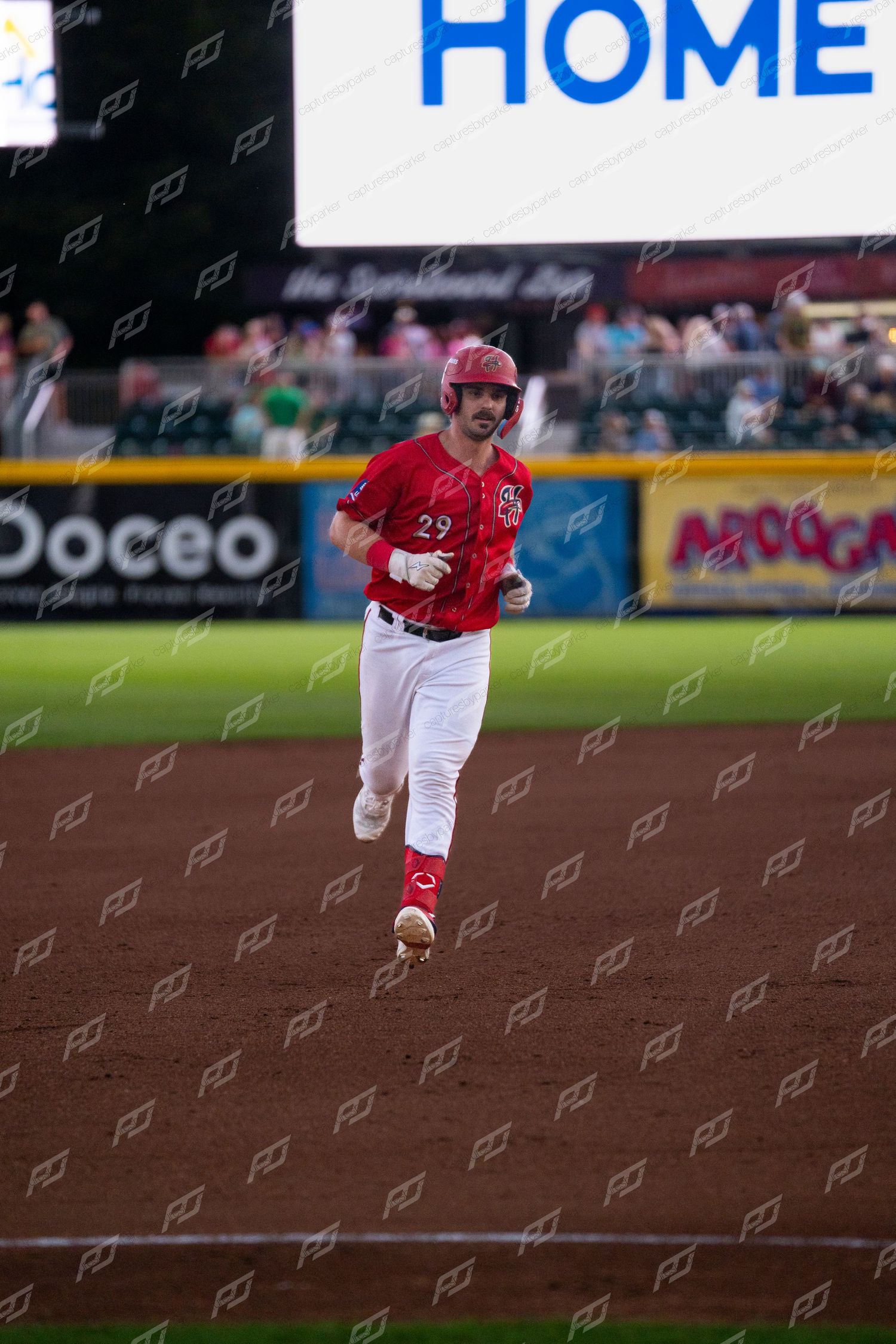 Baseball player #29 in red uniform sprinting the bases during a minor league game under stadium lights.