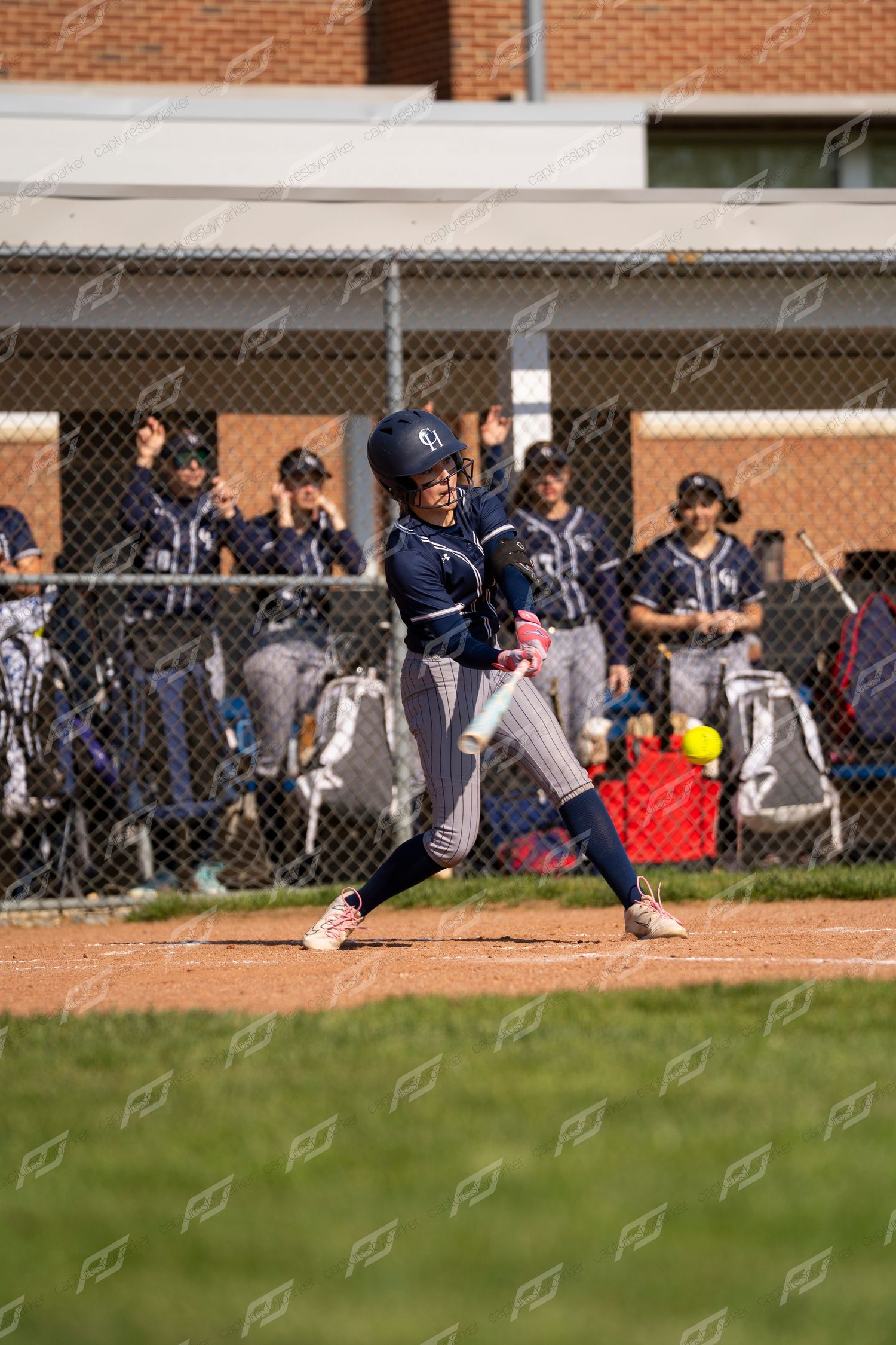 Female softball batter in navy pinstripe uniform swings at yellow pitch during game.