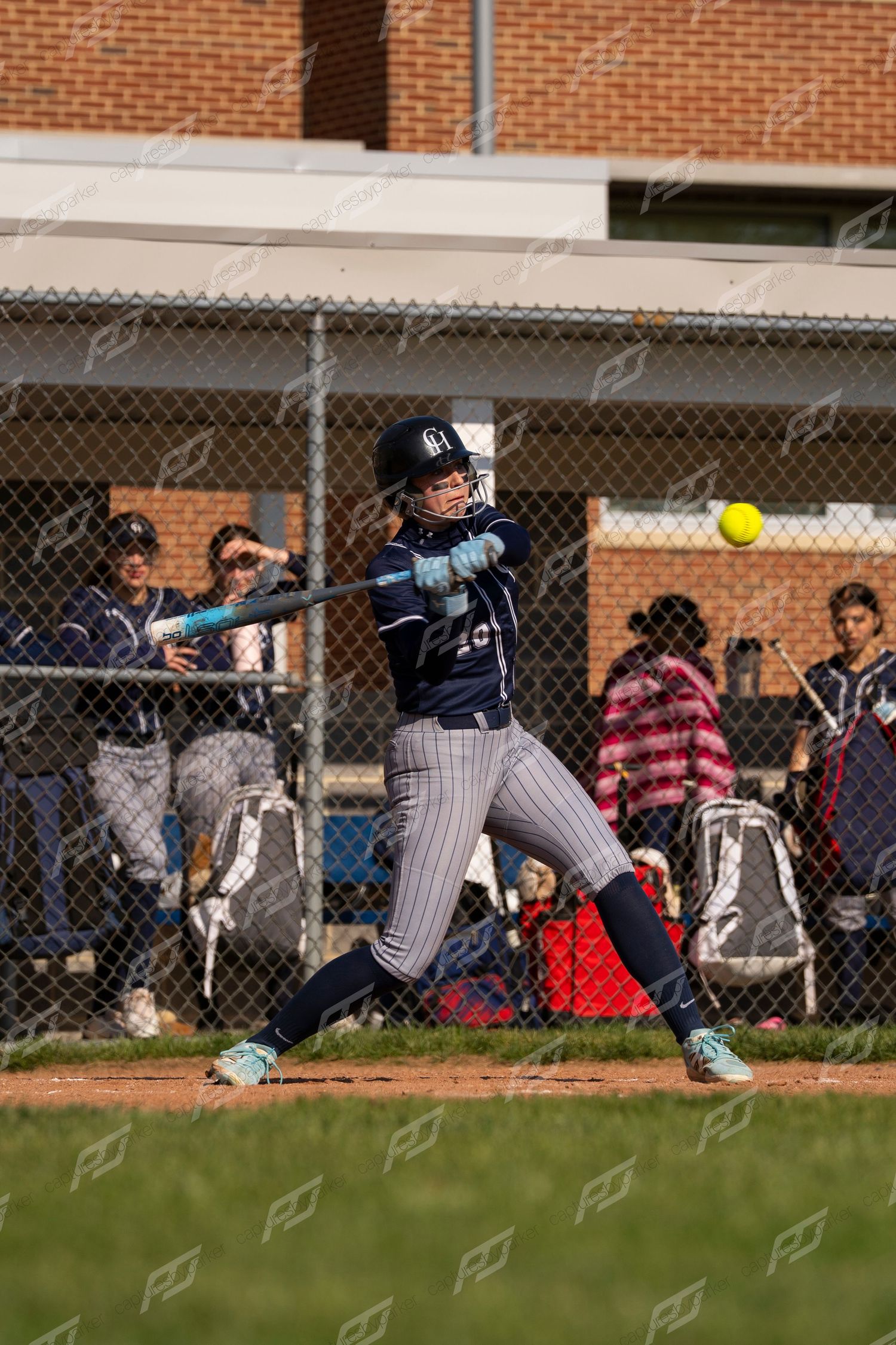 Softball player in navy uniform swinging at a yellow ball during a game with teammates visible in the dugout.