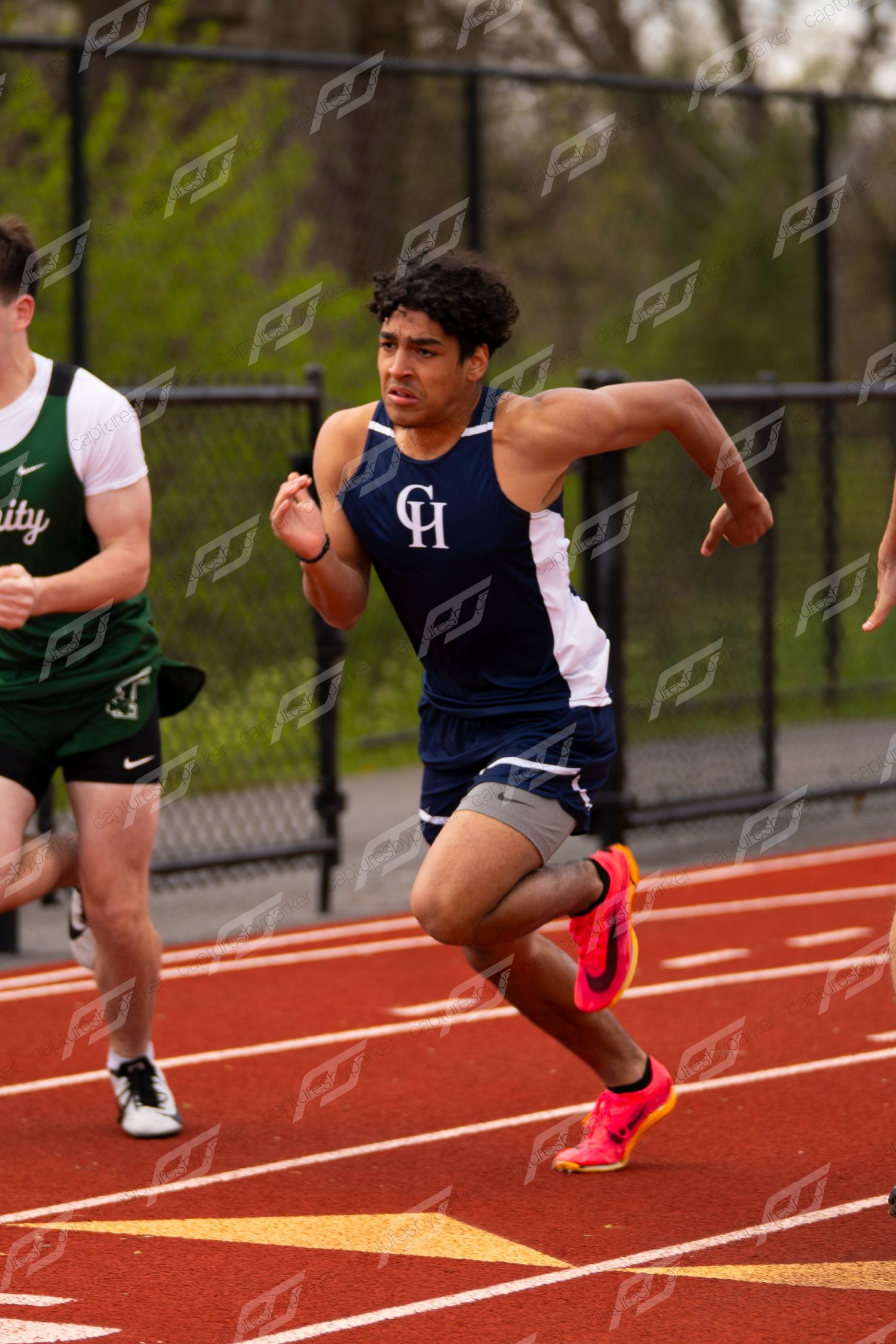Male athlete in CH navy jersey sprints powerfully wearing orange Nike spikes at high school track meet.