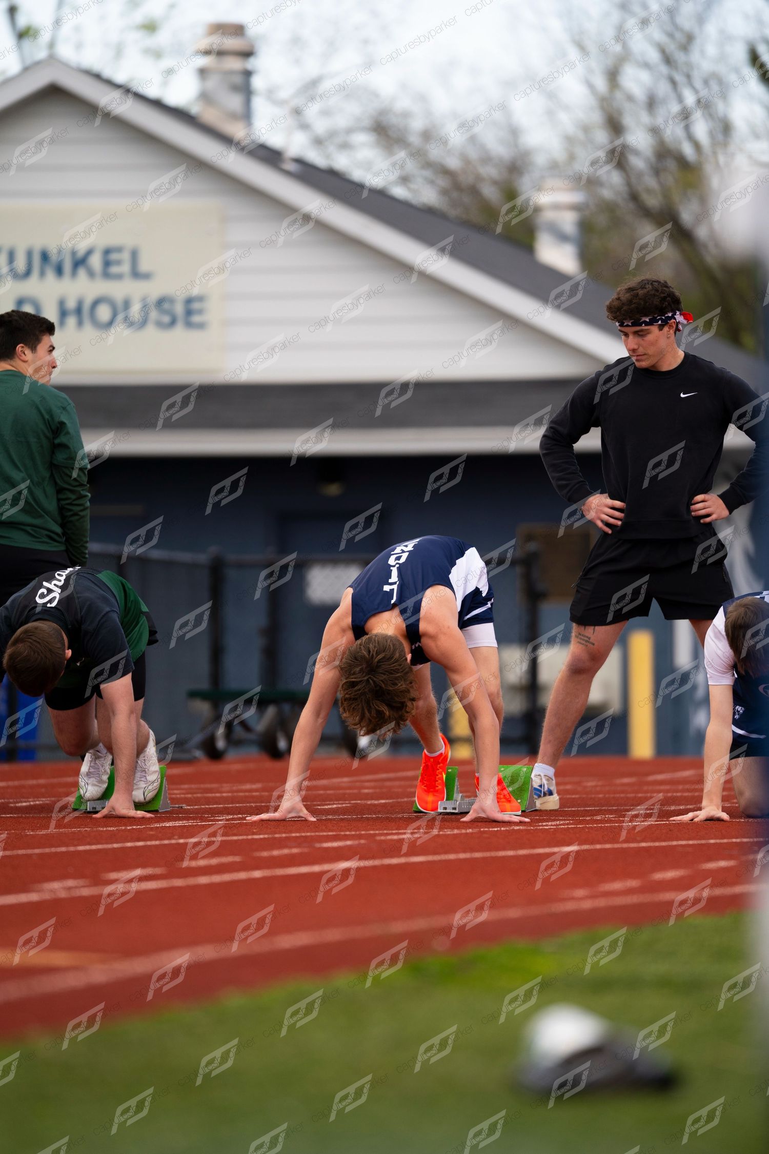 Male sprinter in blue uniform crouches in starting position at blocks on red outdoor track.