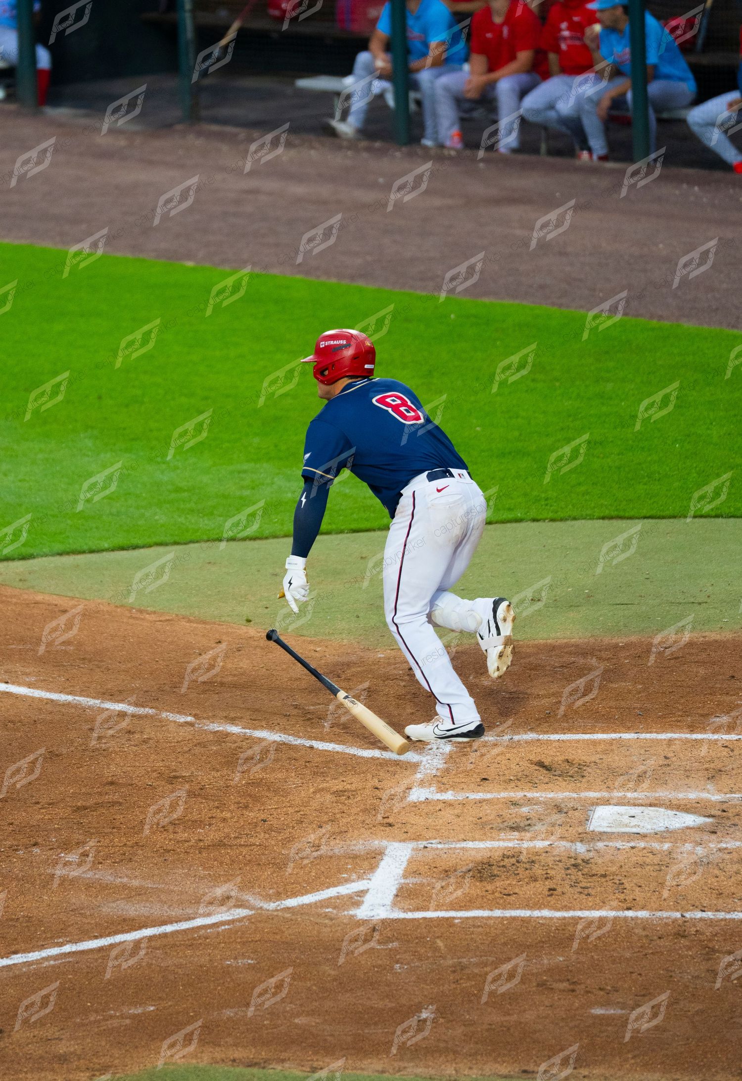 Baseball player in navy uniform drops bat and begins running after making contact during night game.