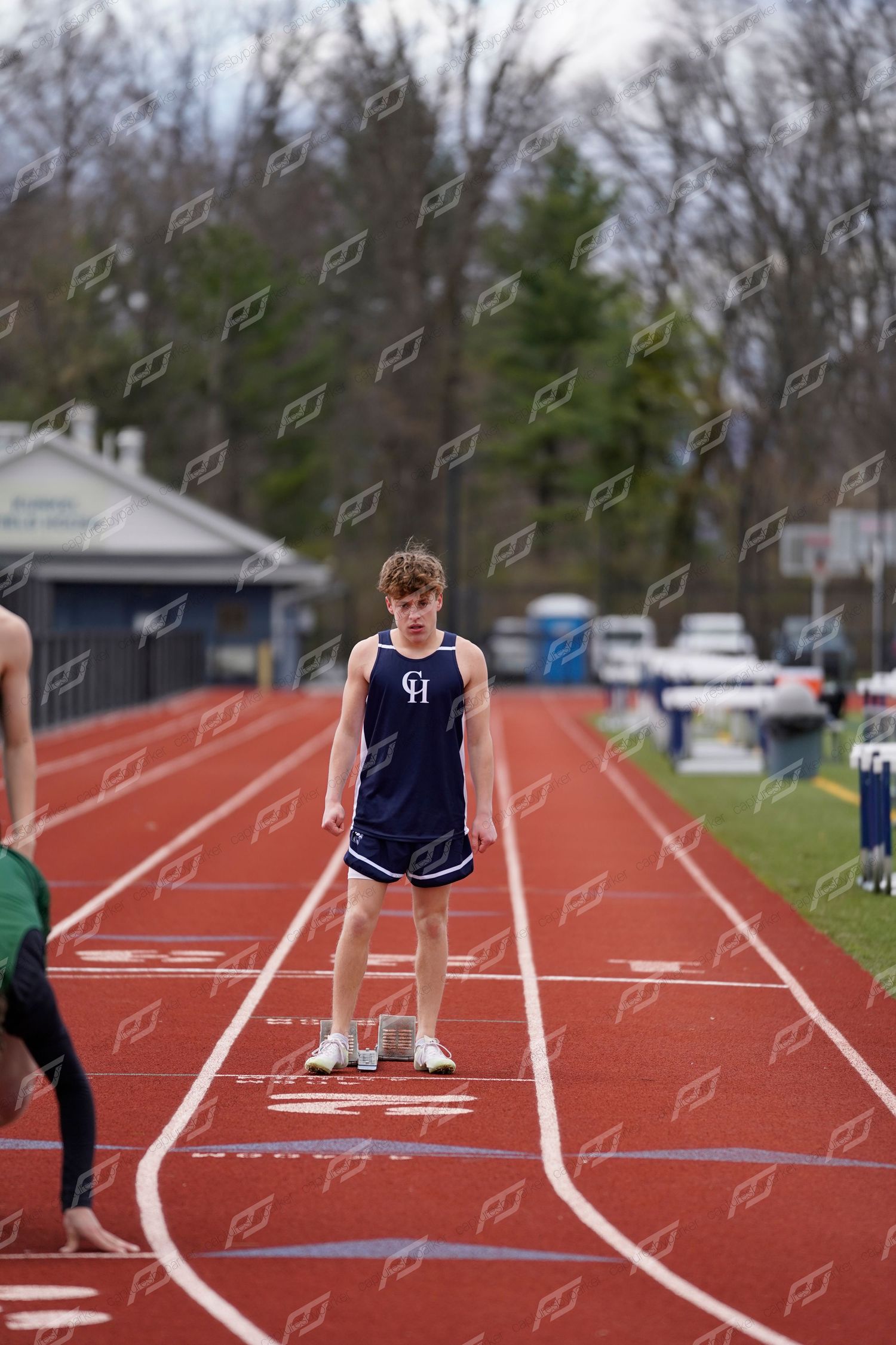 Male track athlete in navy CH uniform stands at starting line on red outdoor track before a race.