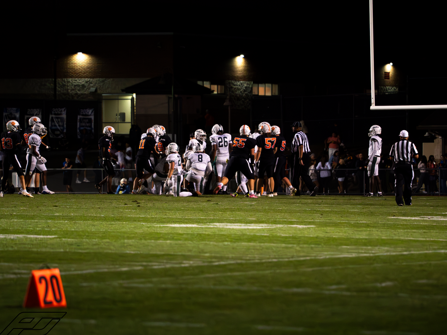 Football teams converge near 20-yard line marker during night game play.