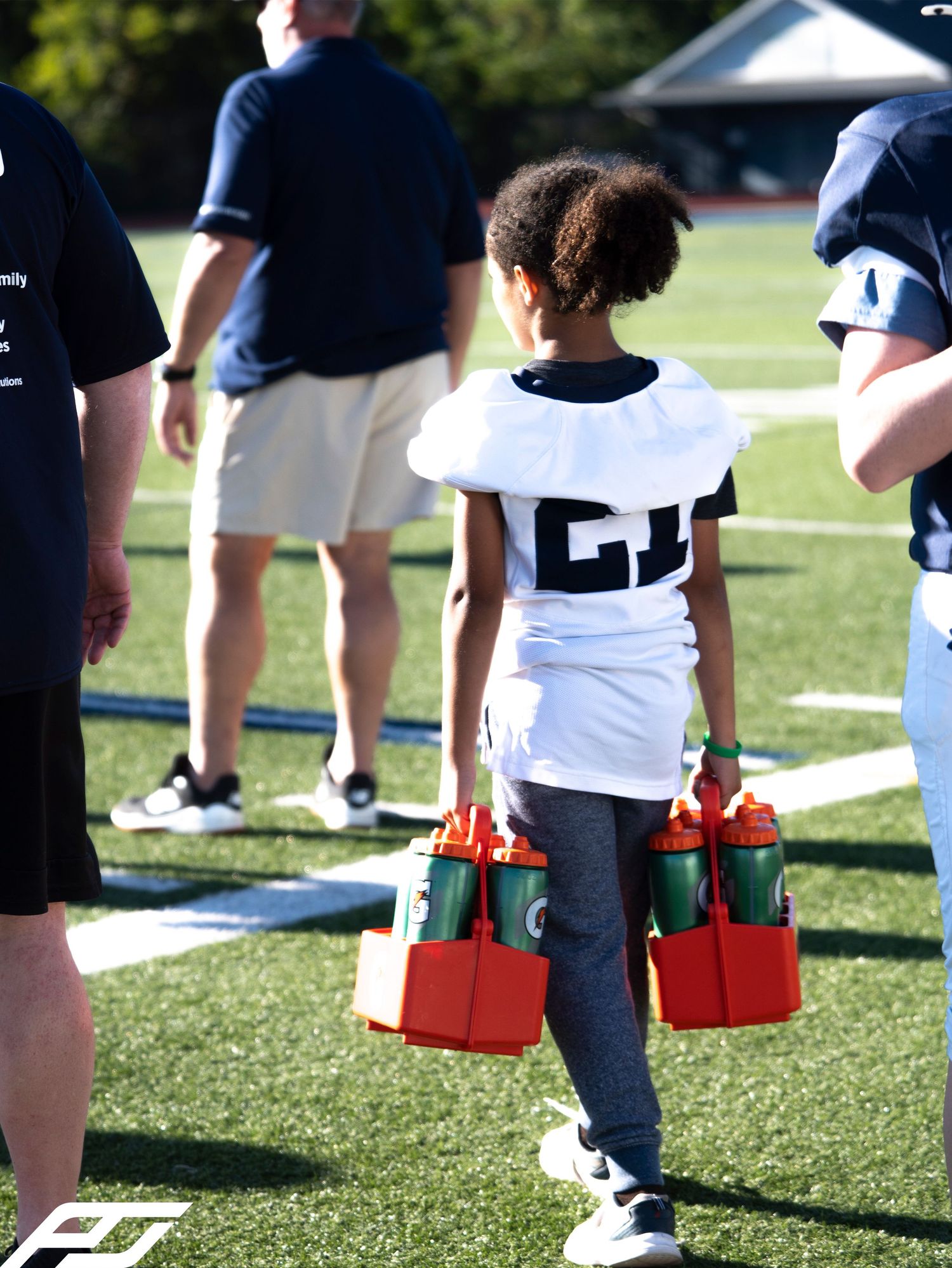 Young football player in jersey #21 carries water bottles on a green field during practice.