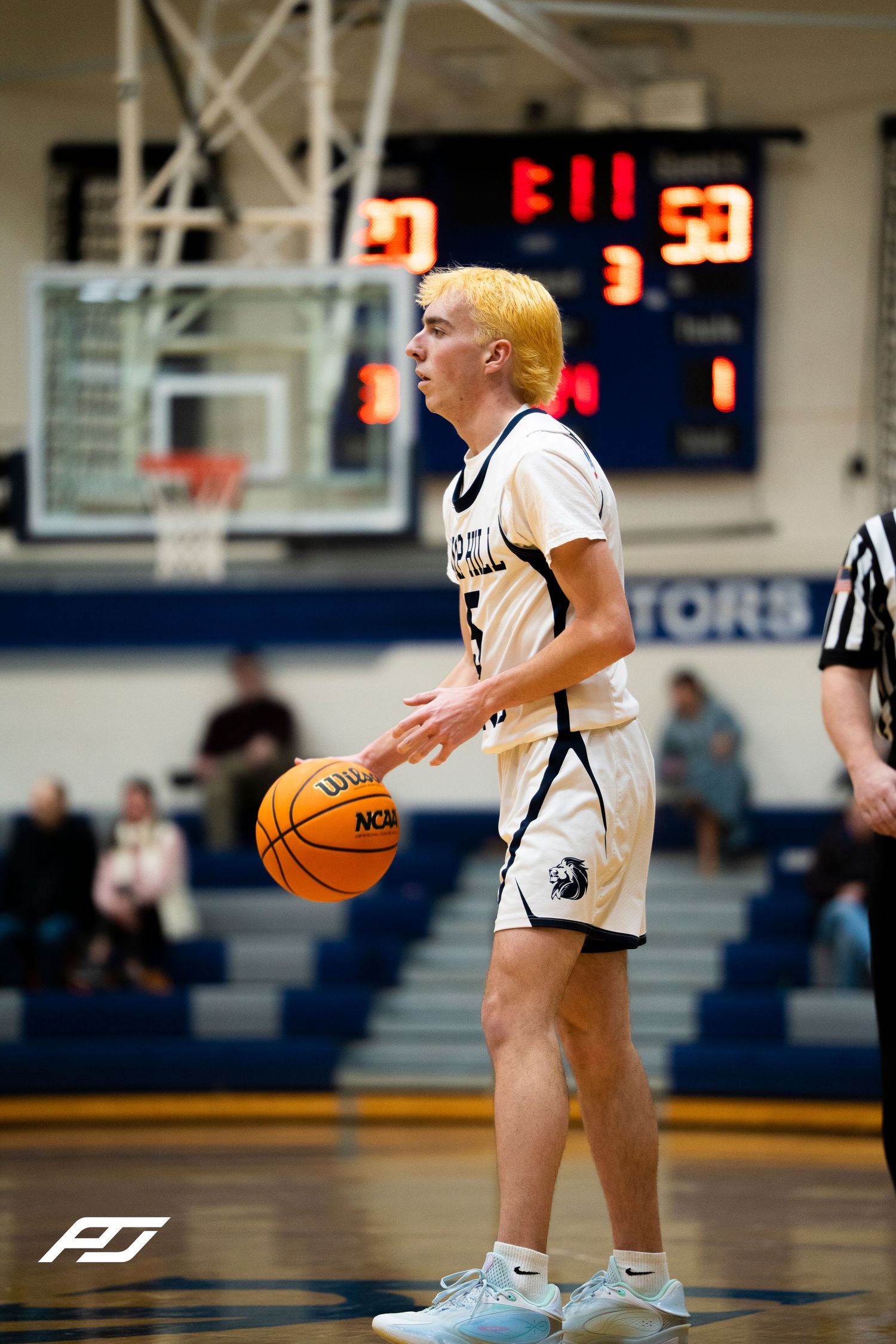 Blonde-haired player #5 in white Lions uniform dribbles basketball during high school game with scoreboard visible.