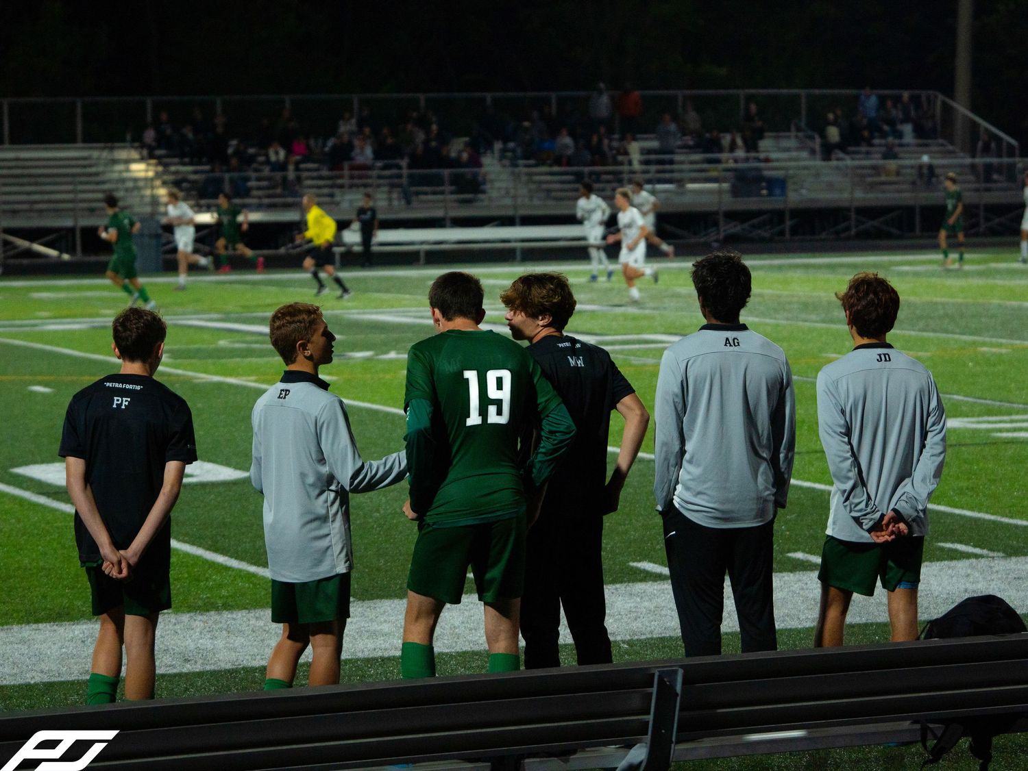 Trinity soccer players in green uniforms watching game action from the sideline at night.
