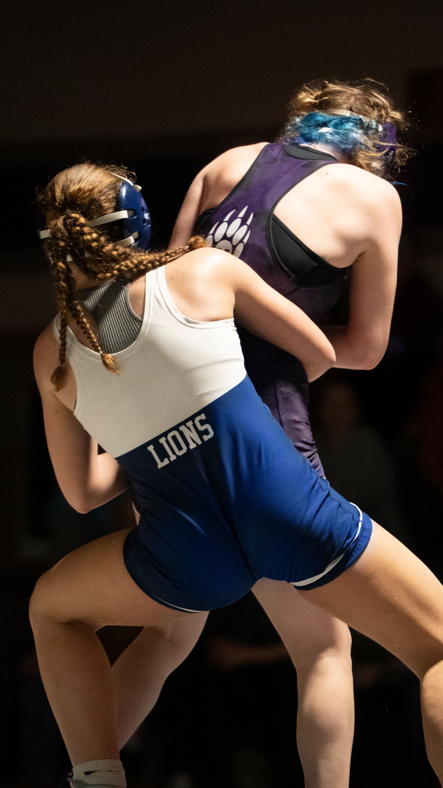 Two female high school wrestlers grapple in a Lions blue singlet versus purple singlet match.