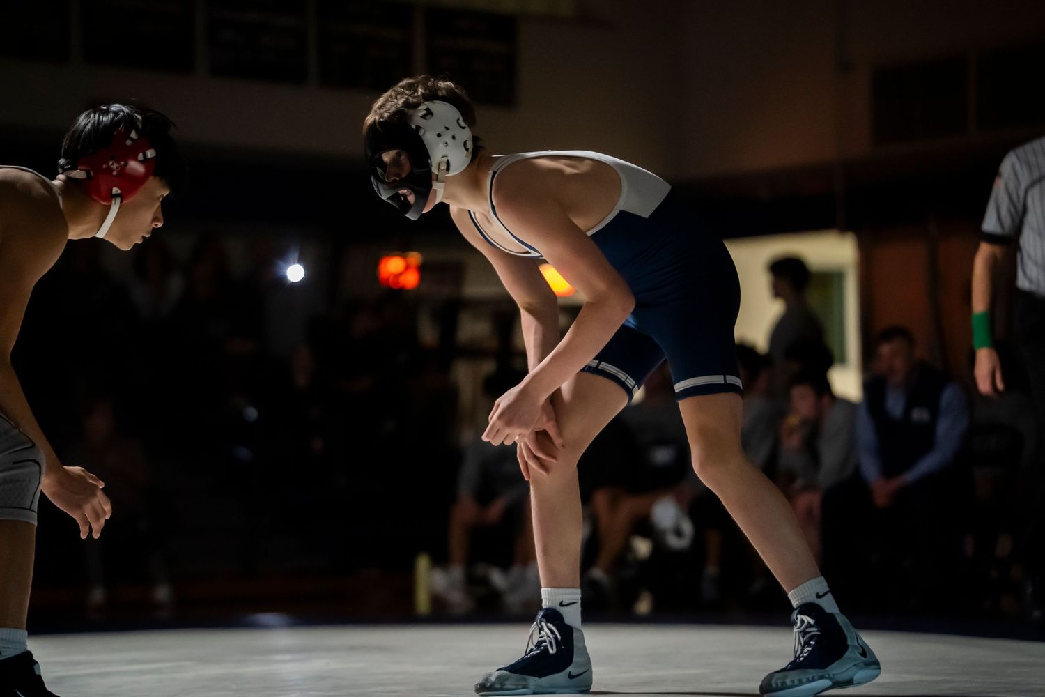 Two high school wrestlers in navy and dark singlets face off in crouched stance on wrestling mat.