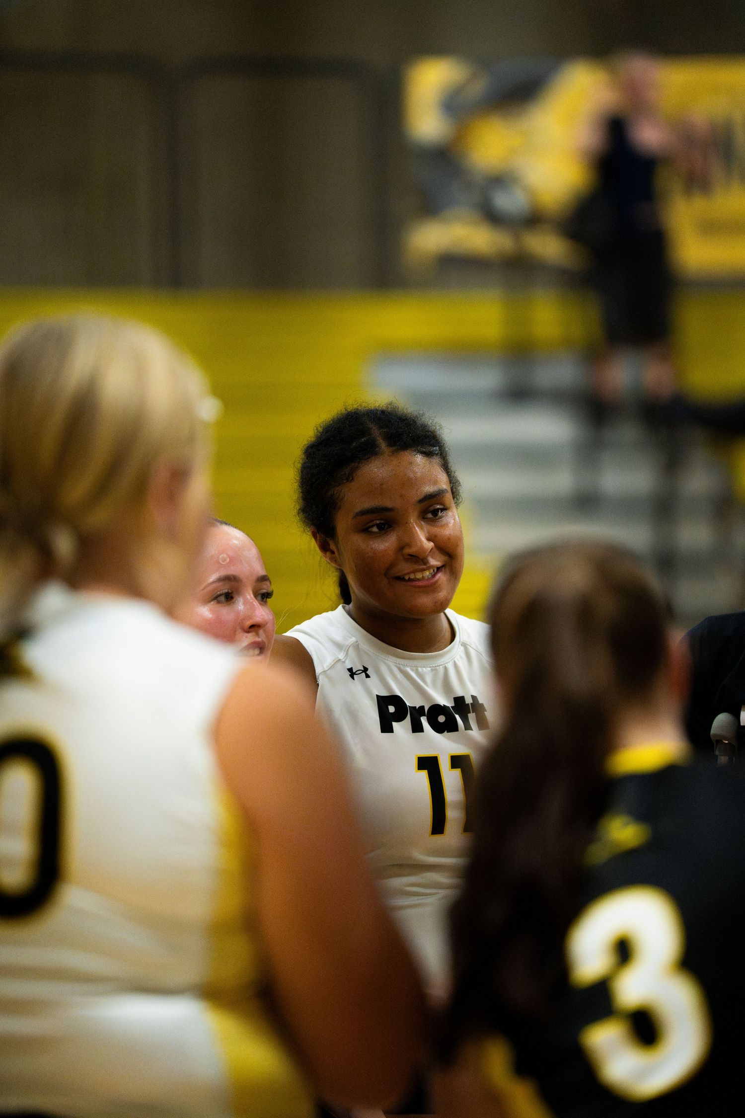 Pratt women's basketball player #11 smiles during a team huddle on the court.