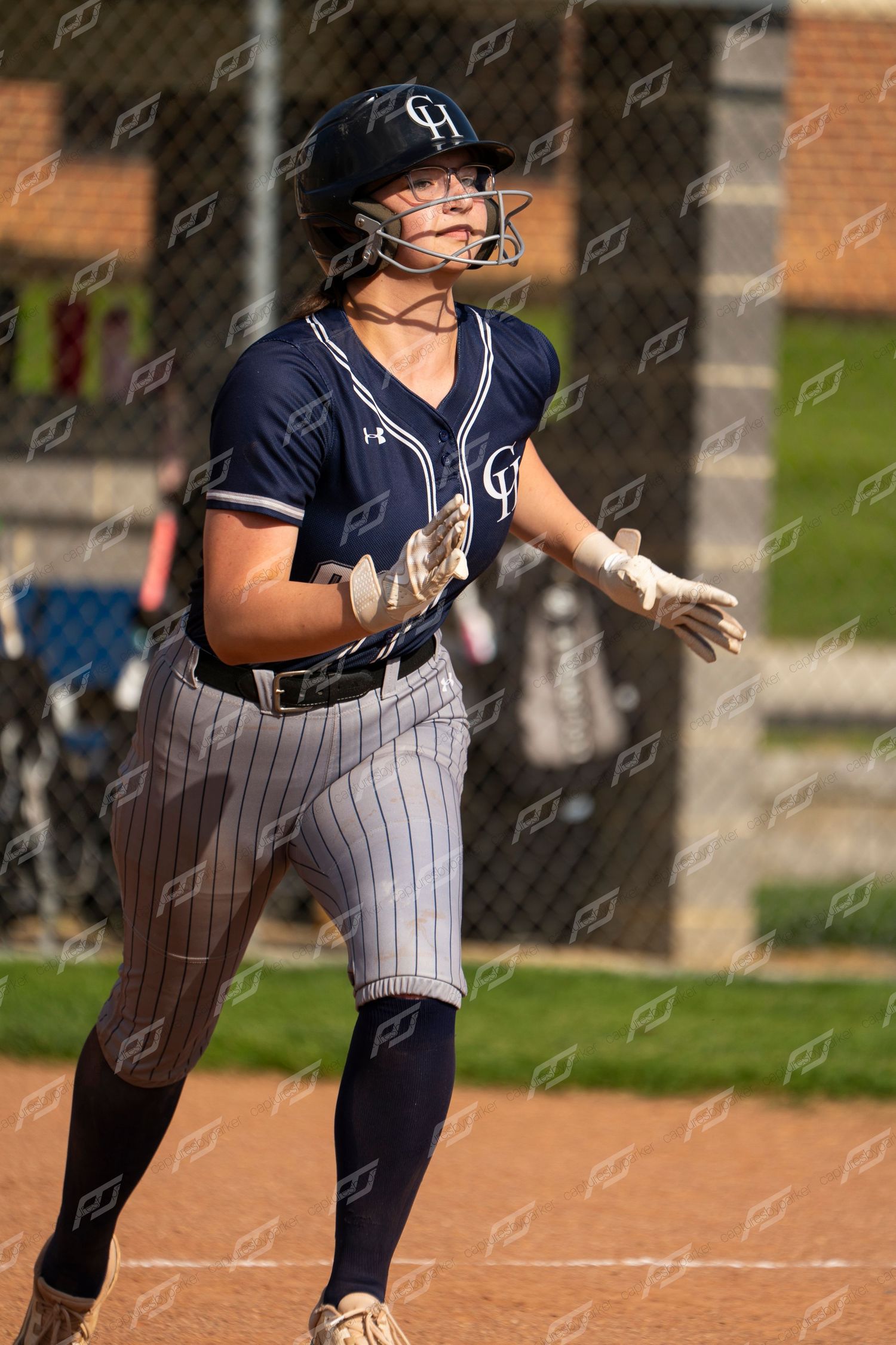 Female softball player in navy CH uniform runs bases while looking up toward sky during game.