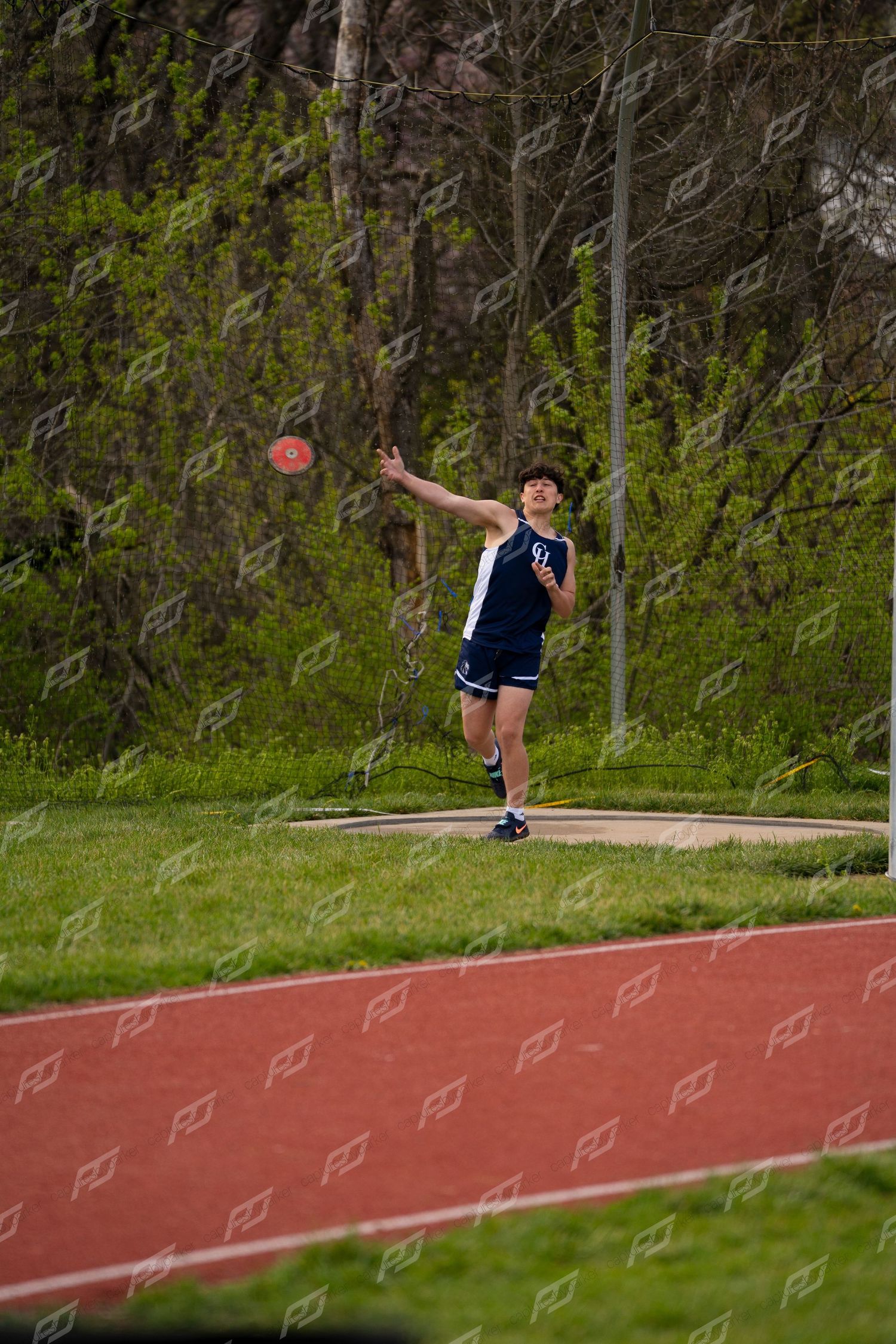Male athlete in navy uniform releases a discus during an outdoor track and field competition.