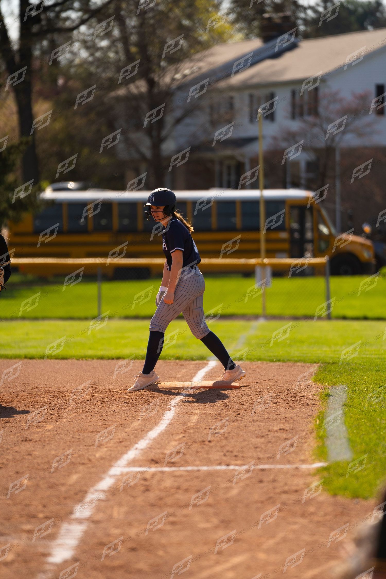 Softball player in navy uniform leads off base during an outdoor game with school bus in background.