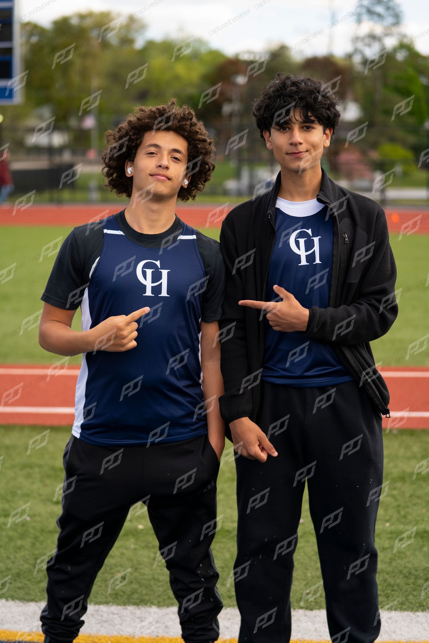 Two young male CH track athletes posing together and pointing at their jerseys on a sports field.