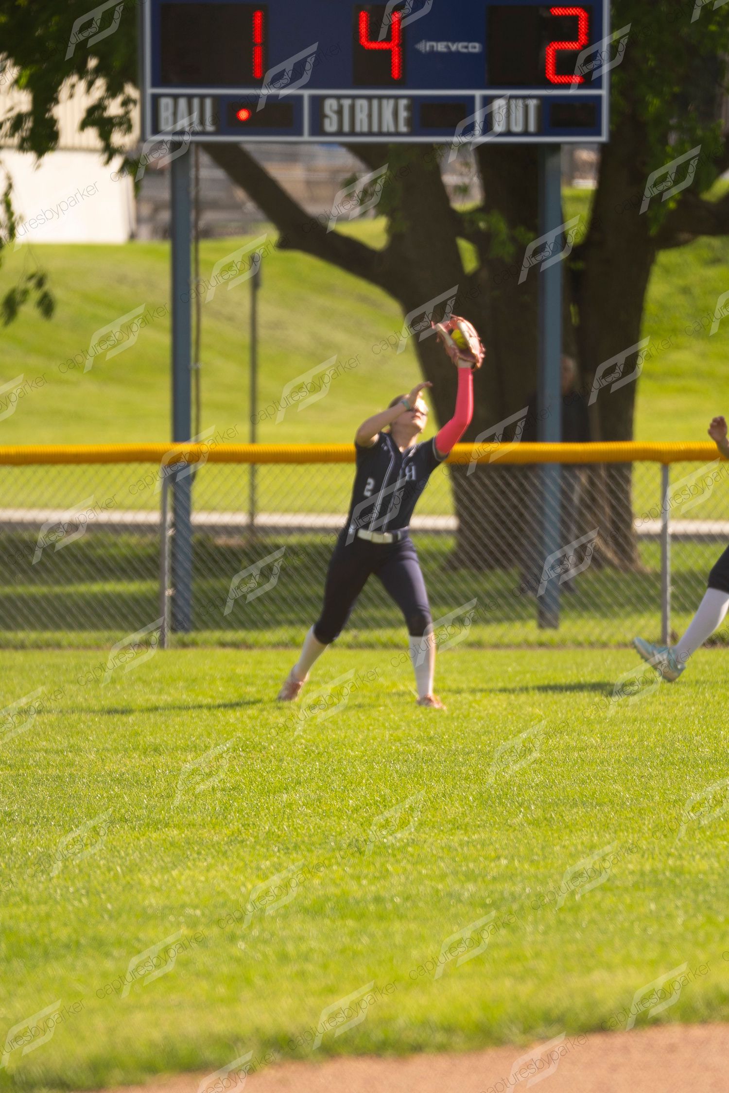 Softball outfielder in navy uniform leaping to catch a fly ball in front of the scoreboard.