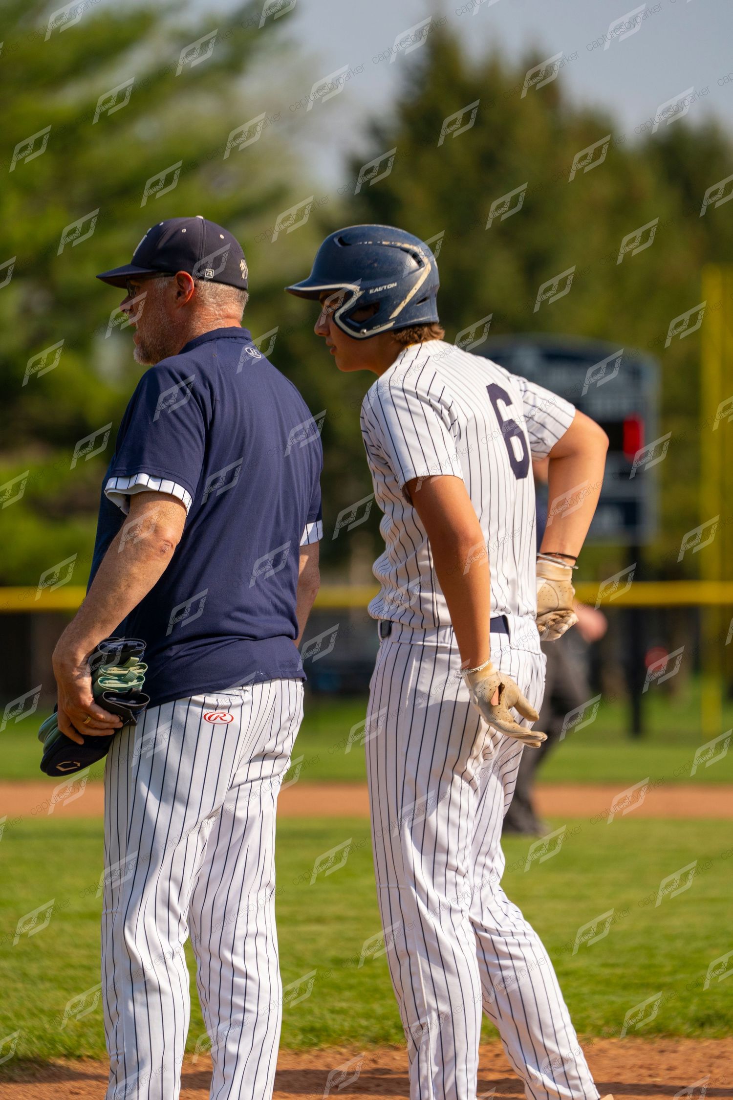 Baseball coach in navy uniform talks with player #6 in white pinstripes at first base.