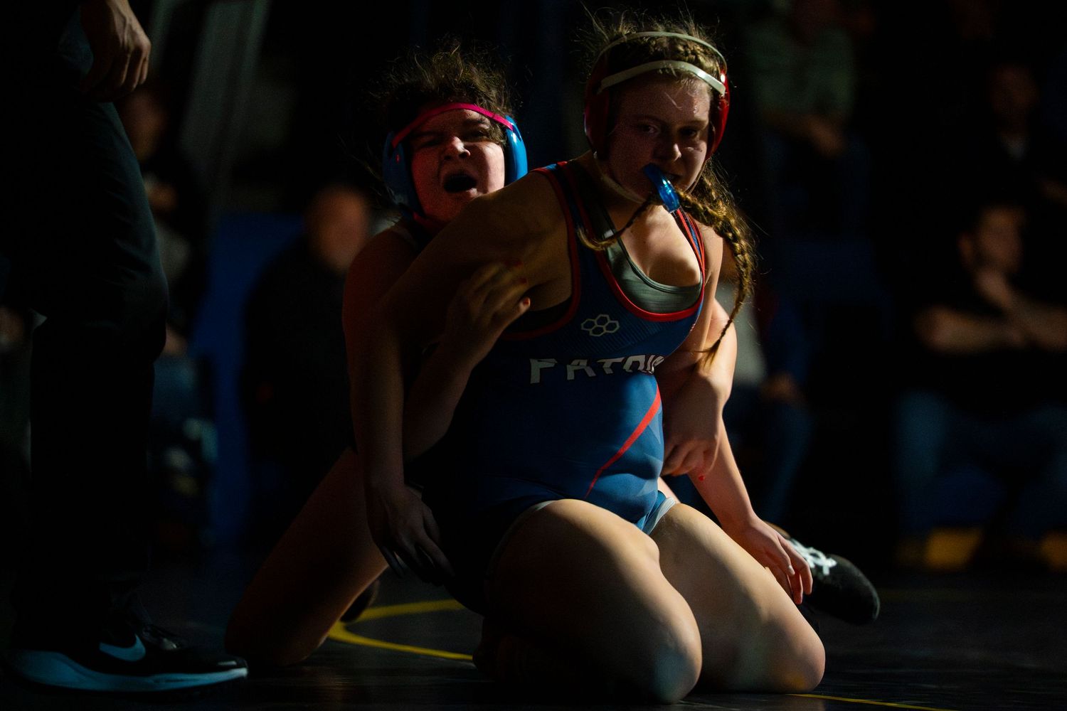 Two female wrestlers compete on the mat, one in a blue singlet controlling her opponent during an intense wrestling match.