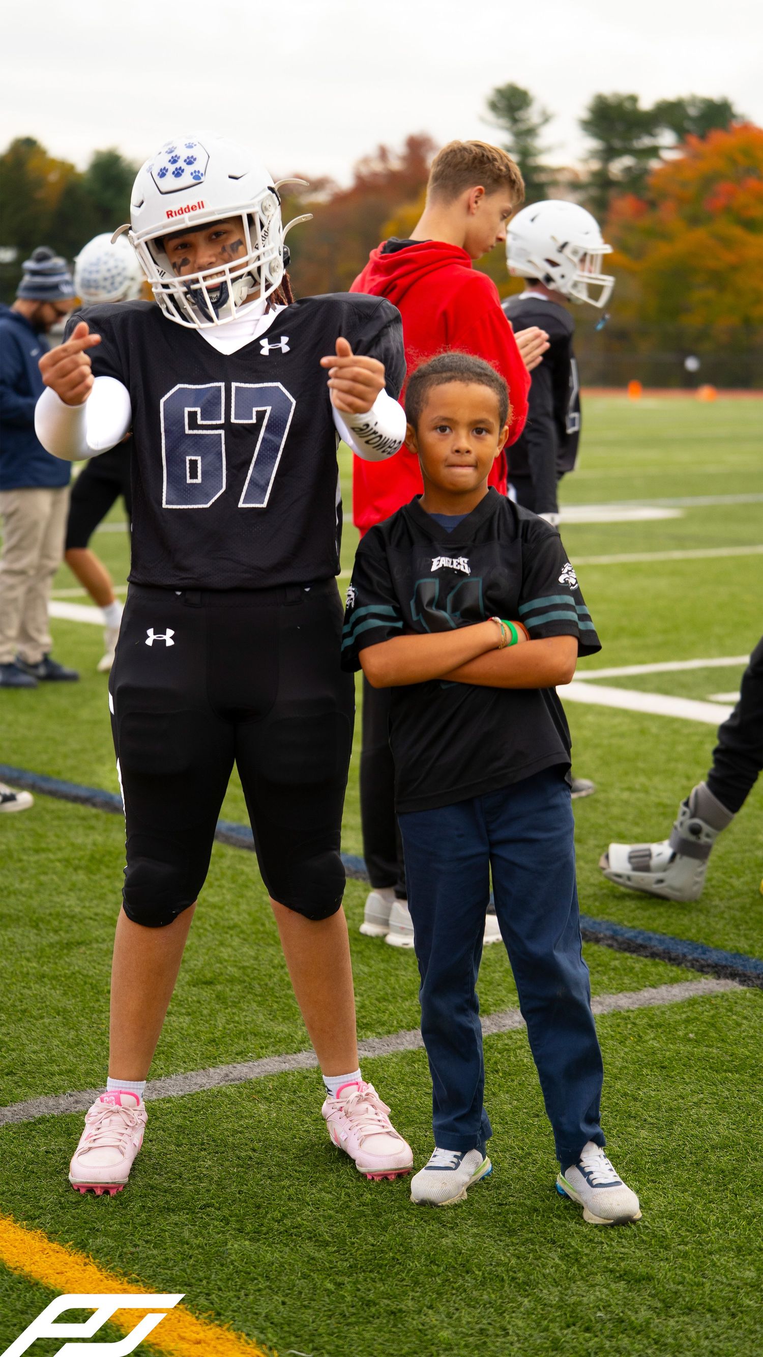 Youth football player number 67 in black uniform gives thumbs up beside a young child on the football field.
