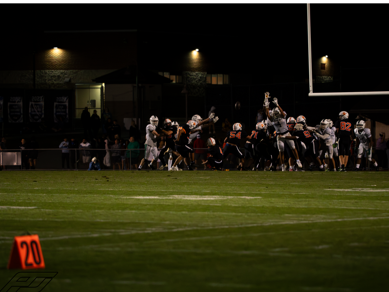 Football players leap and compete near the line of scrimmage under stadium lights with a number 20 yard marker visible.