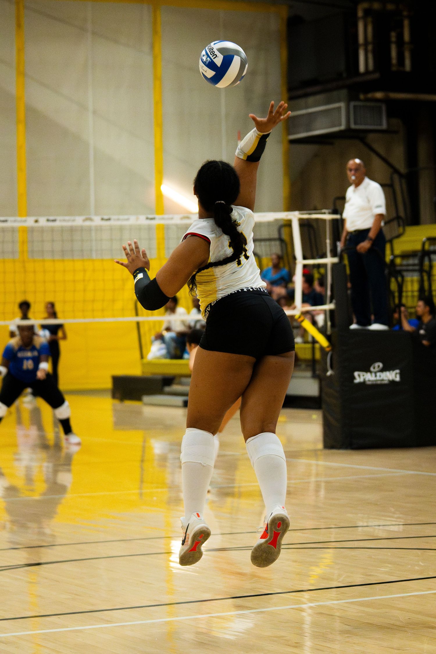 Female volleyball player in black and white uniform jumping to serve ball during indoor match on yellow court.