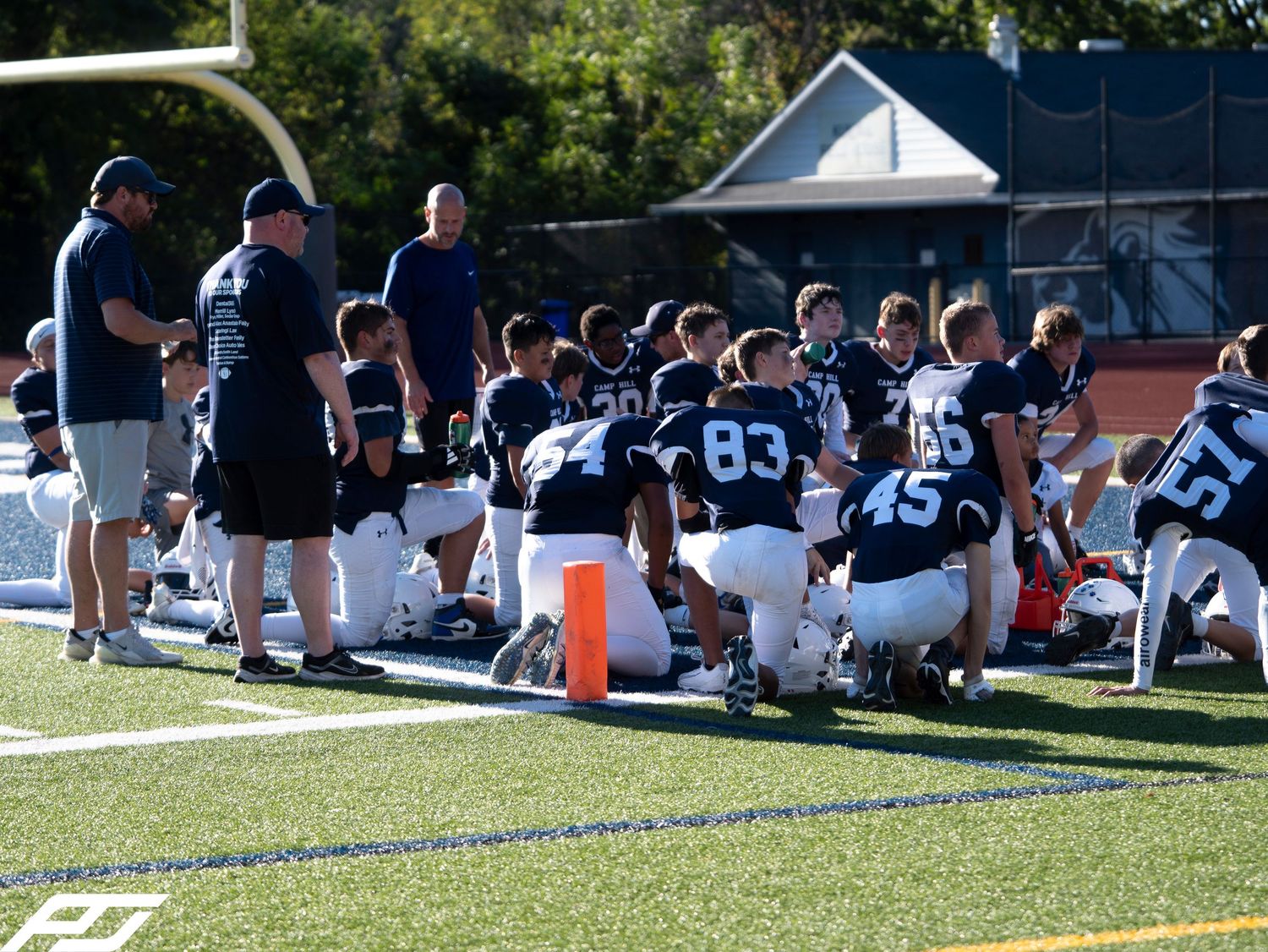 Youth football team in navy uniforms huddles with coaches on the sideline during a game timeout.