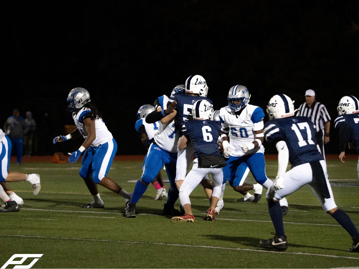 High school football players in navy and blue uniforms clash during night game on lit field.