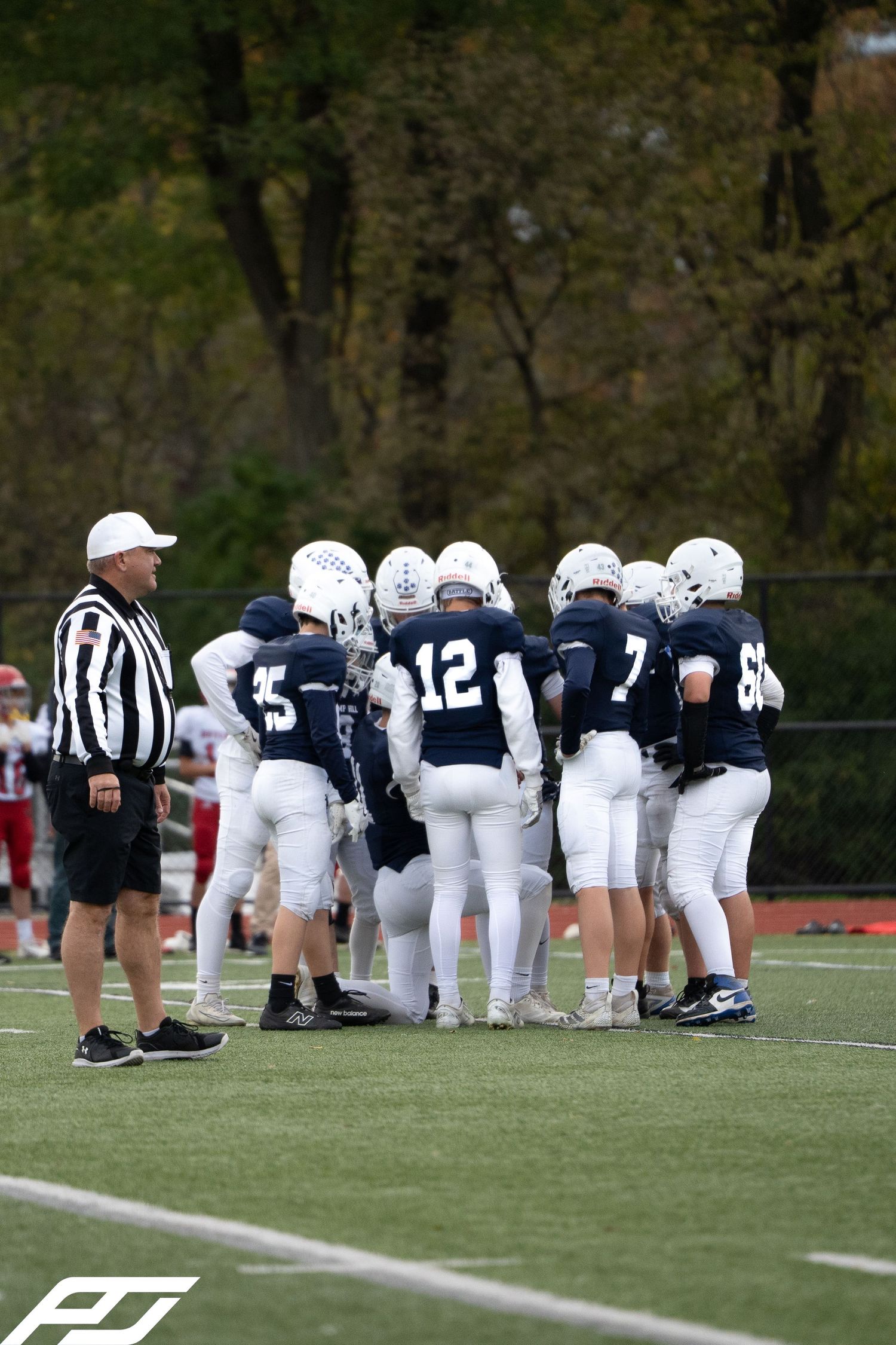 Youth football team in navy uniforms huddles on turf field with coach and referee standing nearby.