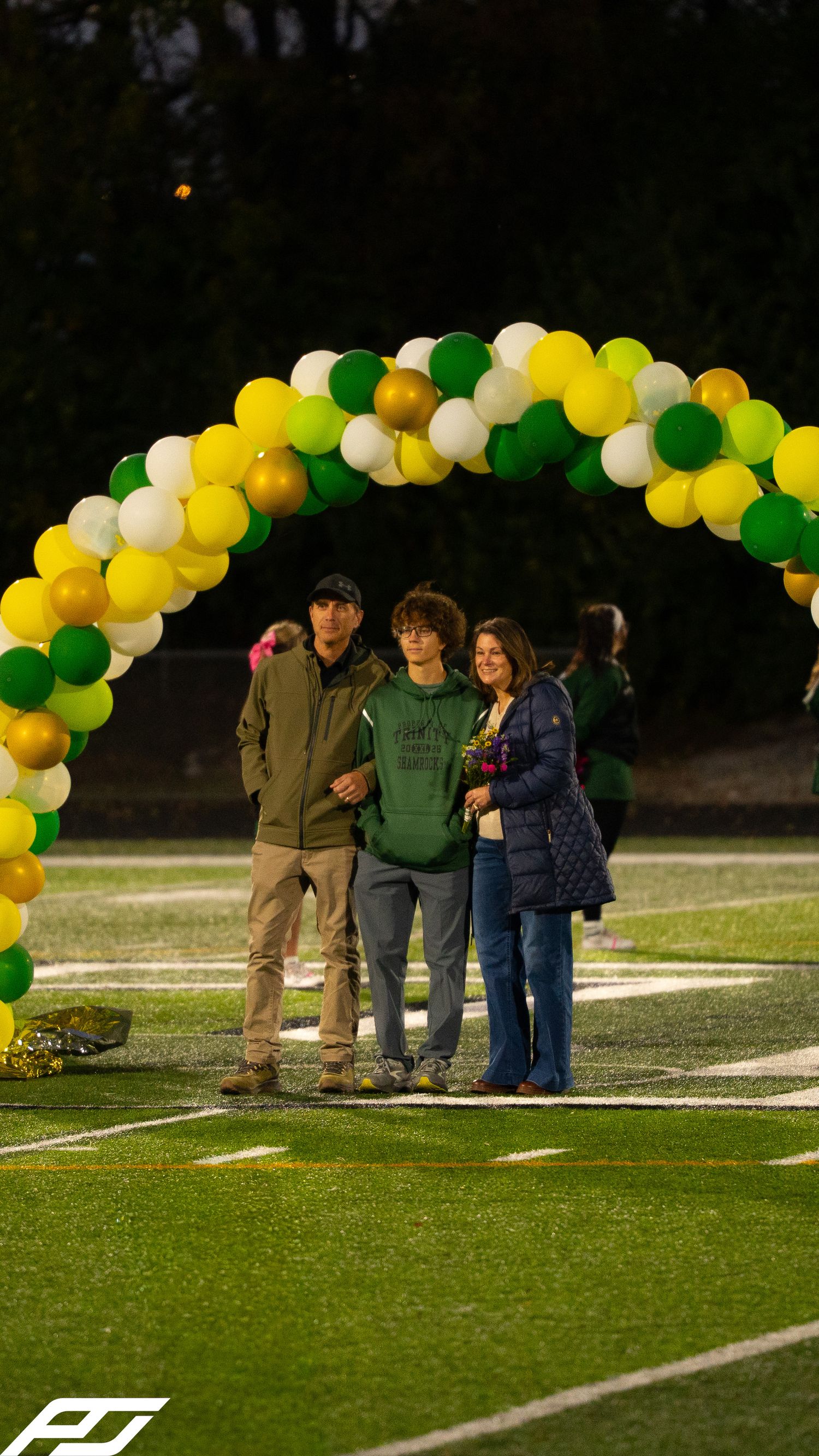 A teenage boy stands with his parents under a green and gold balloon arch during a school senior night ceremony.