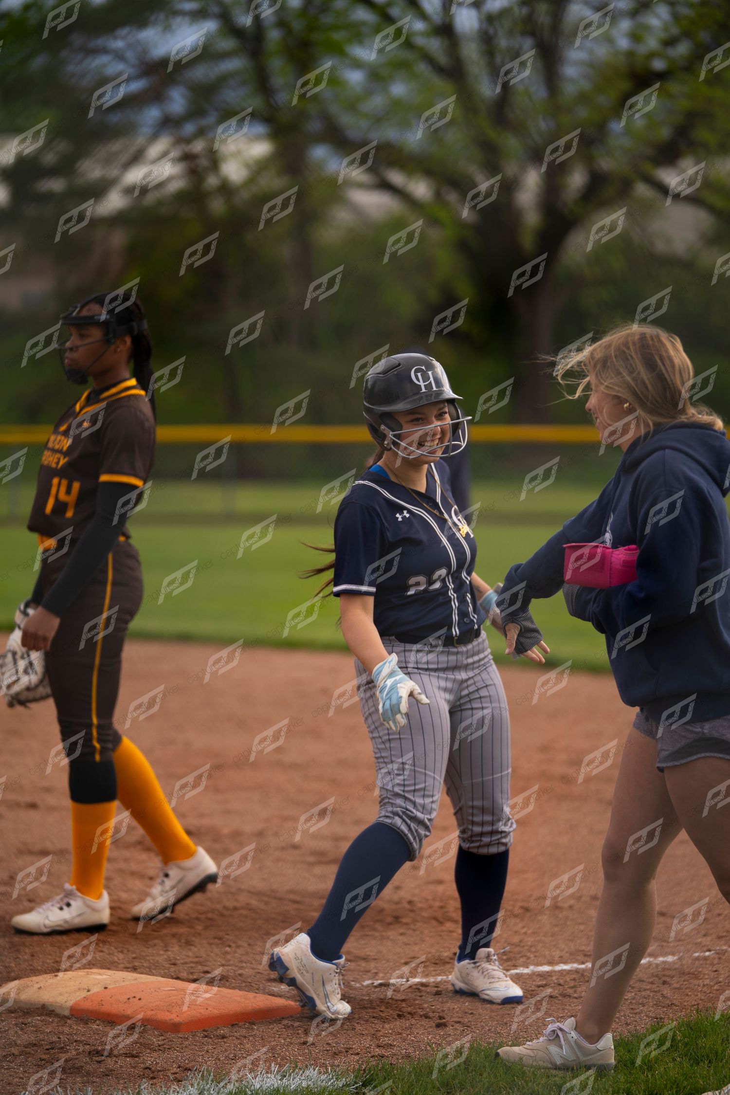 Smiling softball player rounds first base during a game while coach watches nearby.
