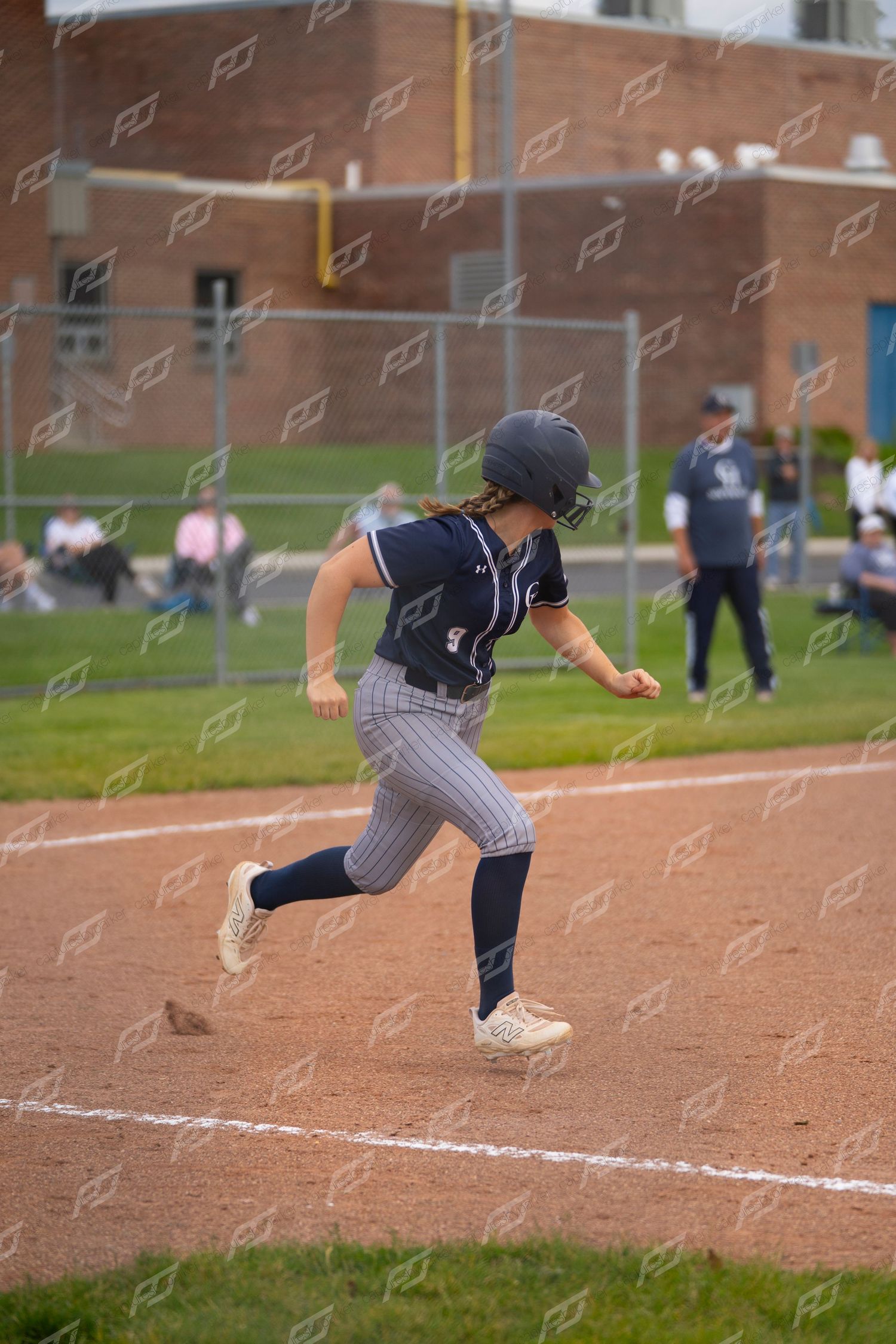 Female softball player in navy uniform and helmet runs the bases during a competitive game.