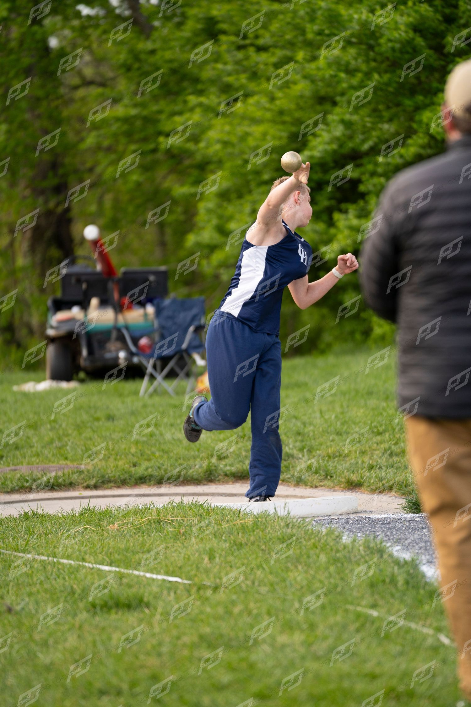 Male athlete releases shot put during outdoor high school track and field competition.