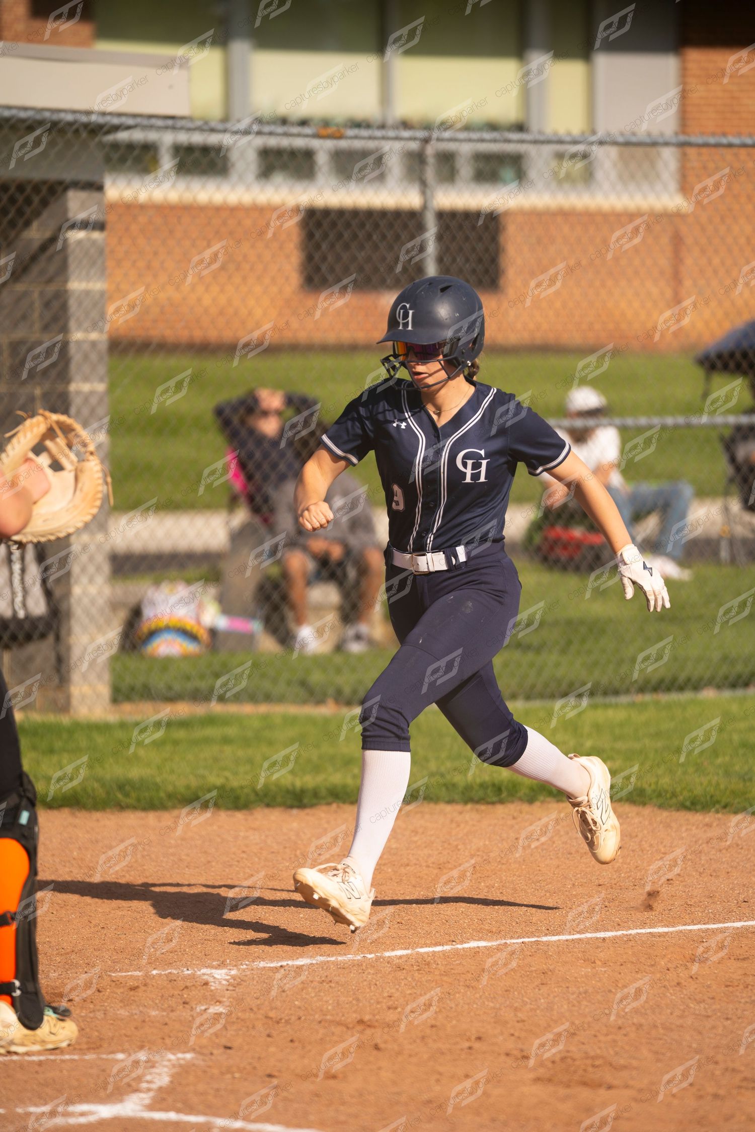 A female softball player in navy CH uniform runs toward home plate during a competitive game.