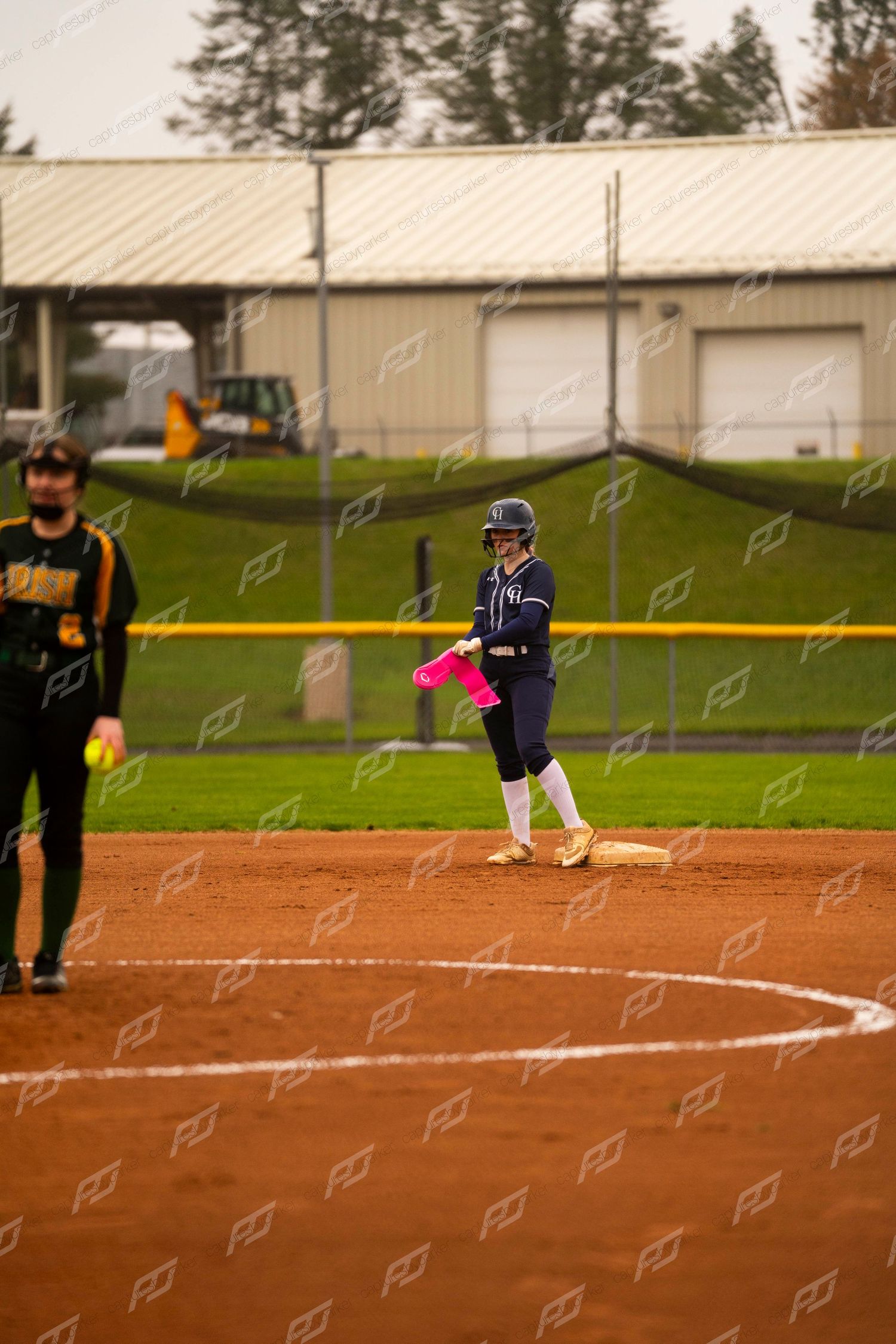 Softball player in navy uniform stands on second base with pink arm sleeve during an overcast game.