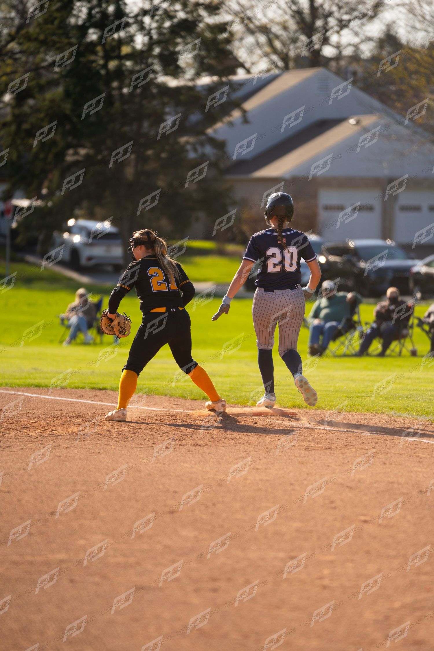 Female softball player #20 in navy uniform running to first base during competitive outdoor game.