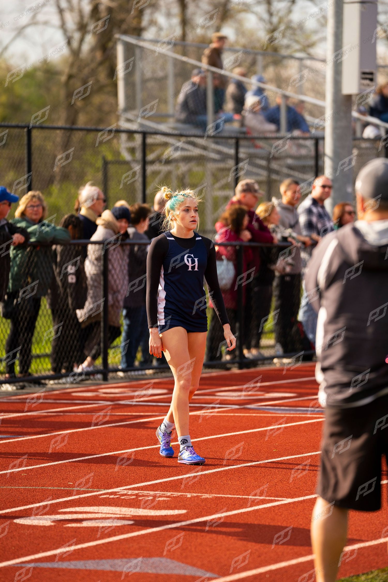 Female high school track athlete in black GH uniform walks on red track before large spectator crowd.